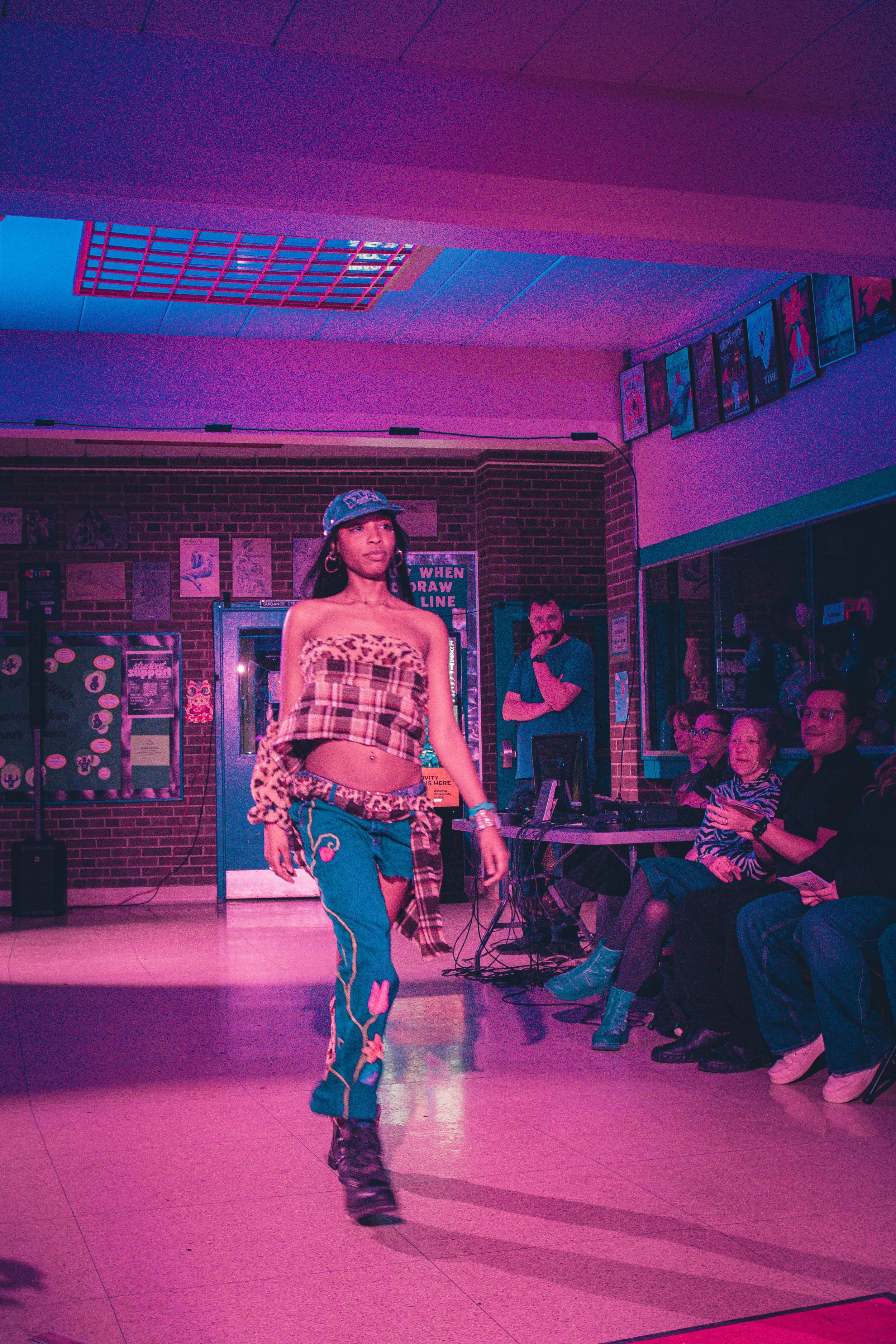 A woman is walking on a stage in front of an audience in a room with brick walls, purple and pink lighting. She is wearing a leopard print tube top, baggy ripped jeans, and a bucket hat. The audience is seated in chairs along the side, watching her a