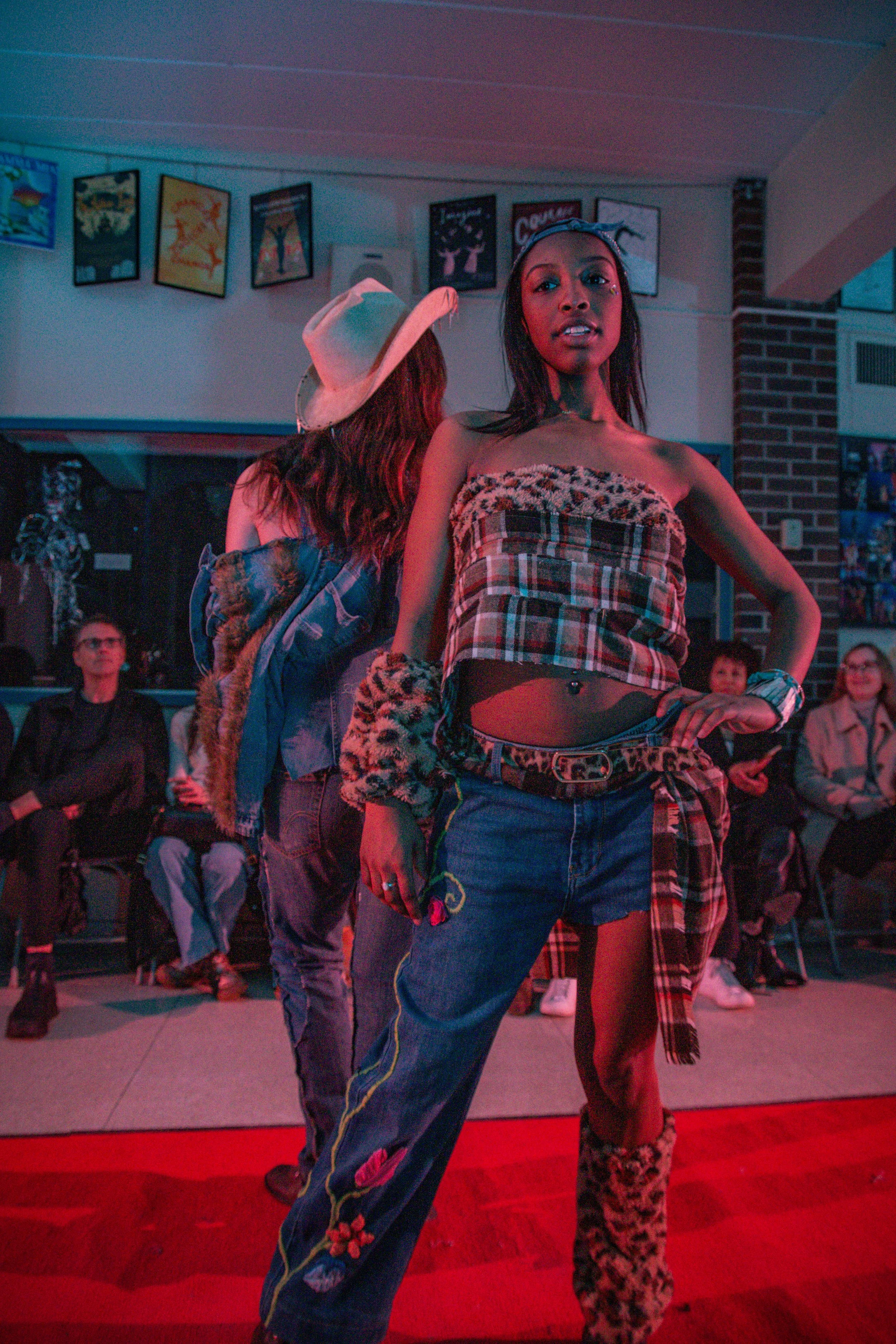 Two women posing in a room with dim lighting. The woman in front has long dark hair, a bandana, and is wearing a plaid tube top, high-waisted jeans, and leopard print leg warmers. The woman behind has long hair and a hat, wearing a denim jacket with 