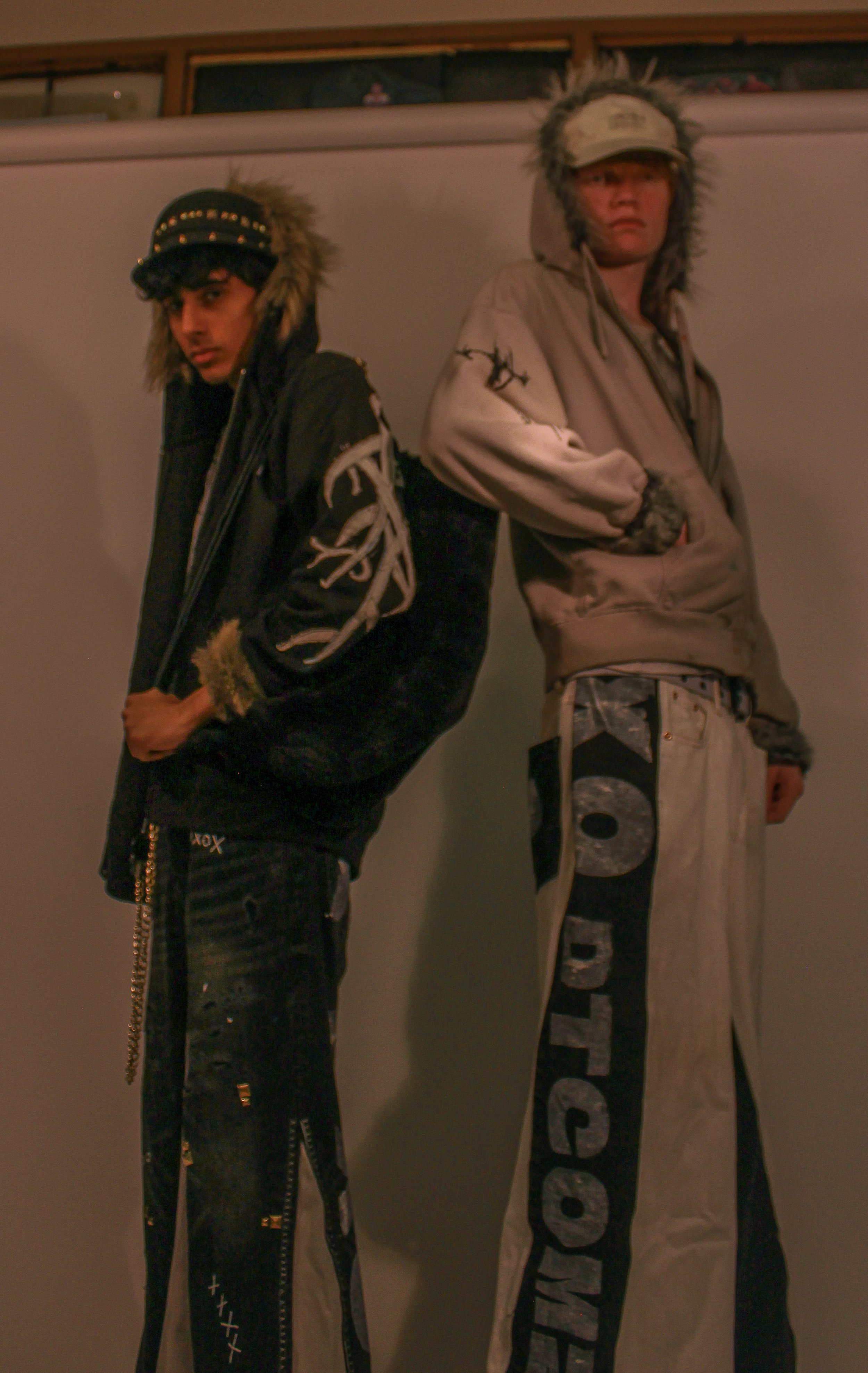 Two young men standing against a wall, wearing hooded jackets with faux fur trim and black pants with writing or designs on them.