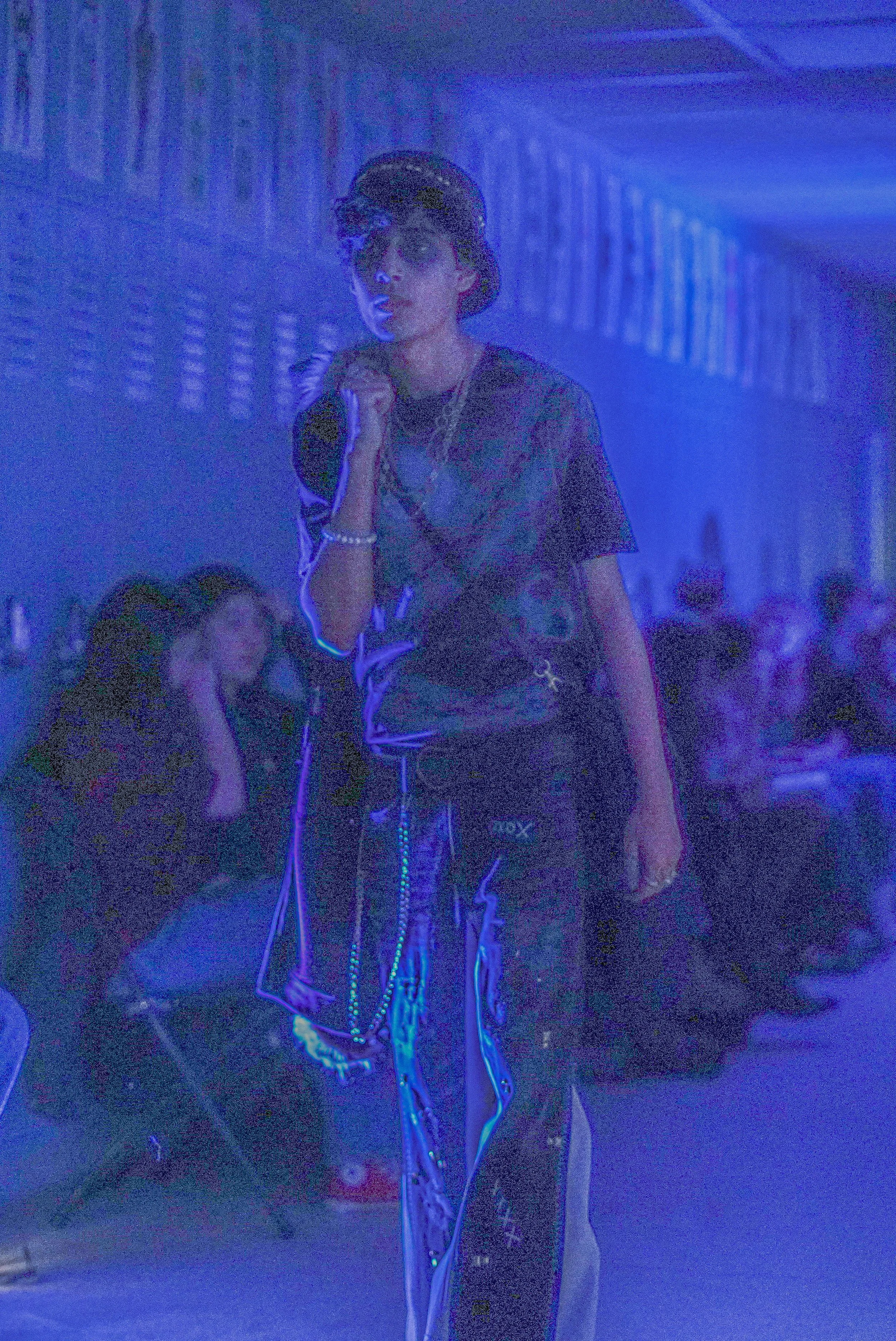 A person dressed in streetwear with chains, glasses, and a cap, walking in a dimly lit indoor space with blue lighting and other people in the background.