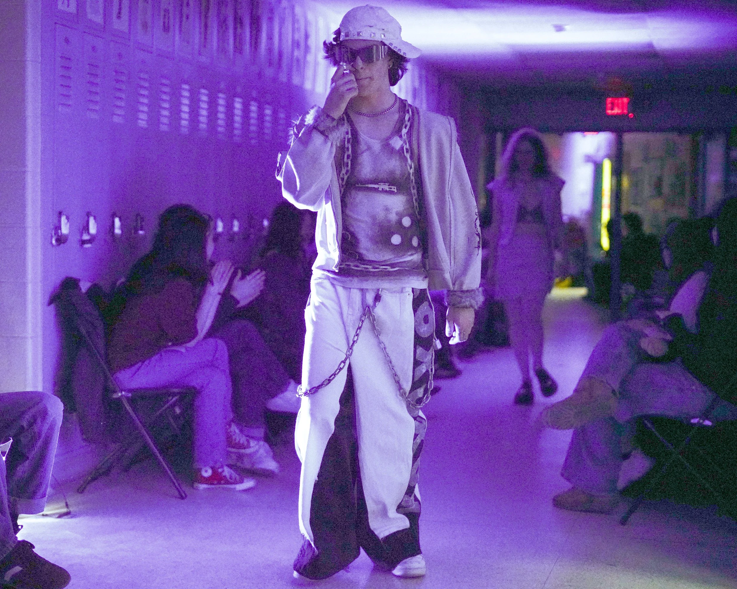 A young person in a white cap and large sunglasses looking at their phone, wearing stylish chains and a jacket, standing in a dimly lit hallway with purple lighting, and people sitting or standing in the background.