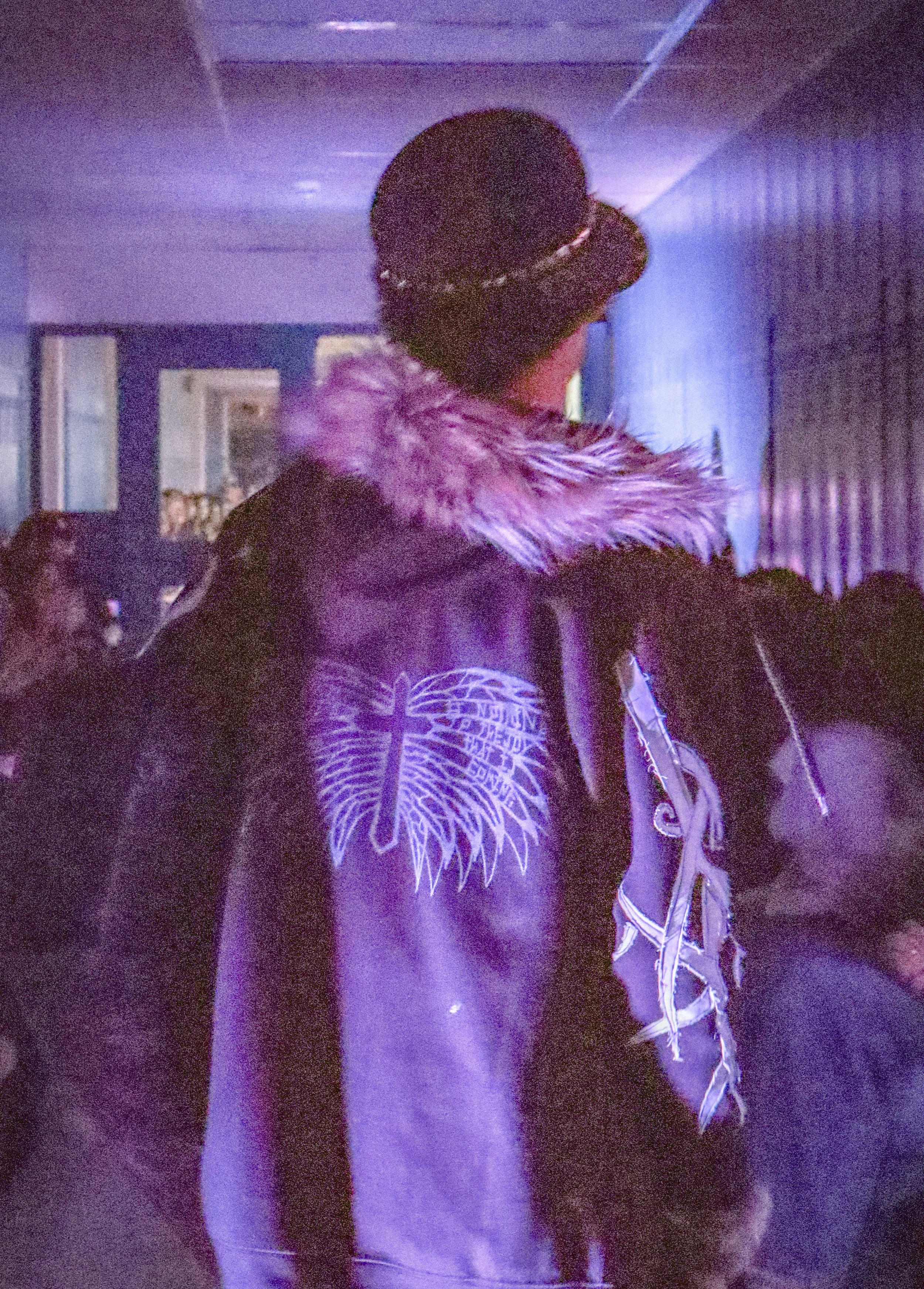 A person wearing a black hoodie with a white graphic design on the front, a black baseball cap with a pattern, and a wolf fur-lined jacket in a dimly lit, purple-toned setting.