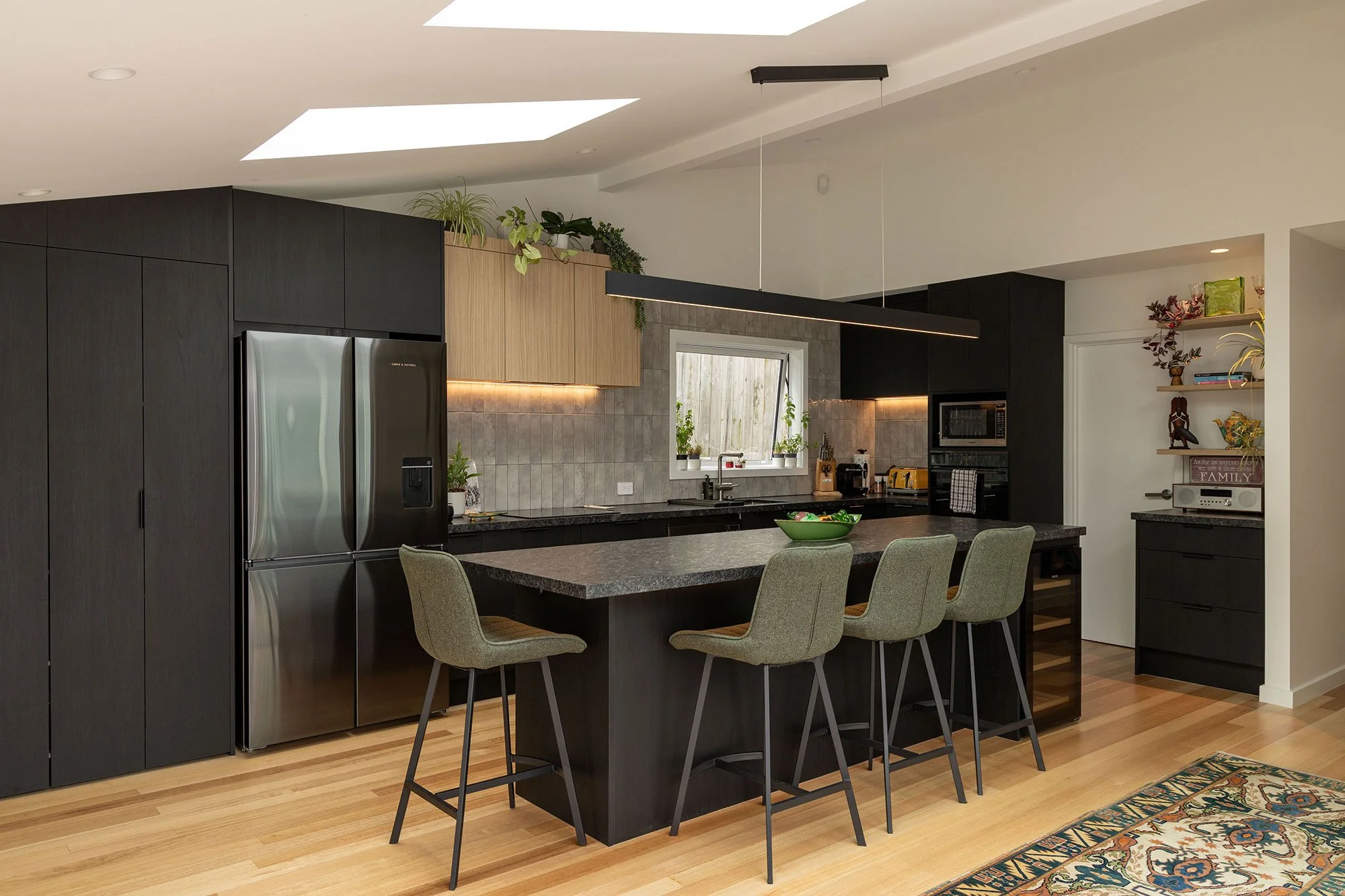 Renovated modern kitchen extension in Auckland with black cabinets, stainless steel refrigerator, gray backsplash, and a black island with beige chairs. There are plants on top of cabinets and sunlight coming through a window.