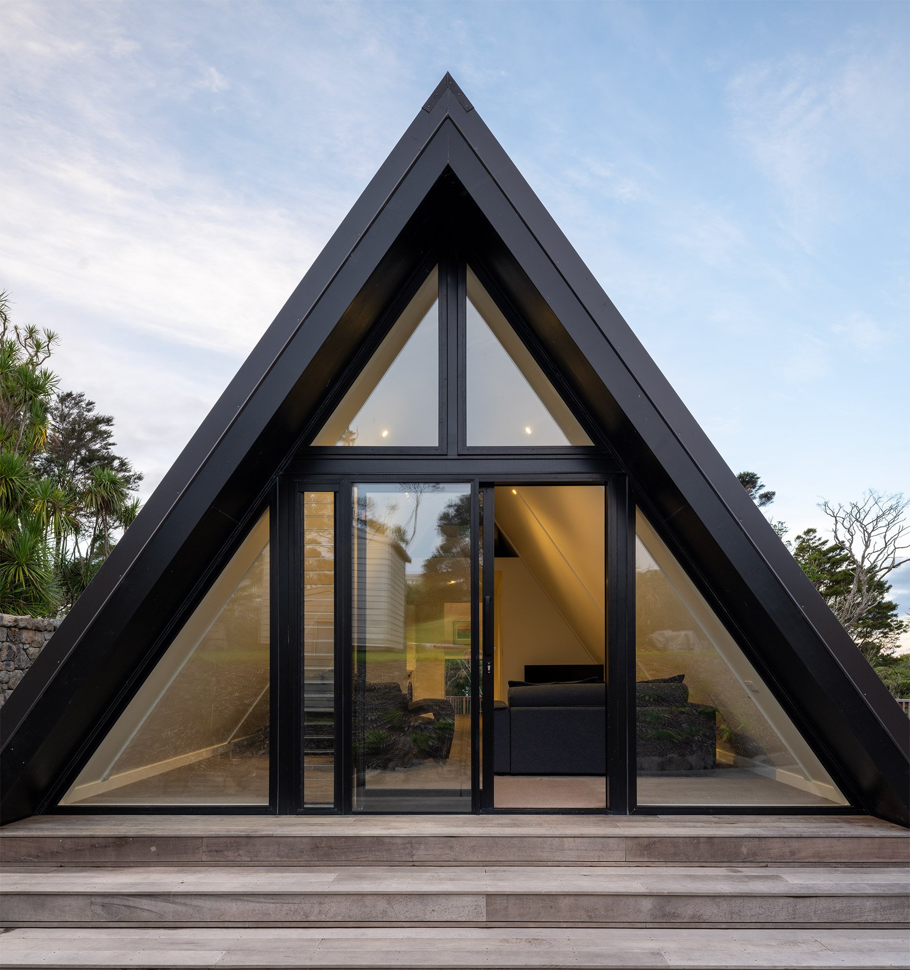 A modern new build A-frame house in Auckland with large glass doors and windows, black exterior, and a wooden deck with steps, set outdoors with trees and sky in background.