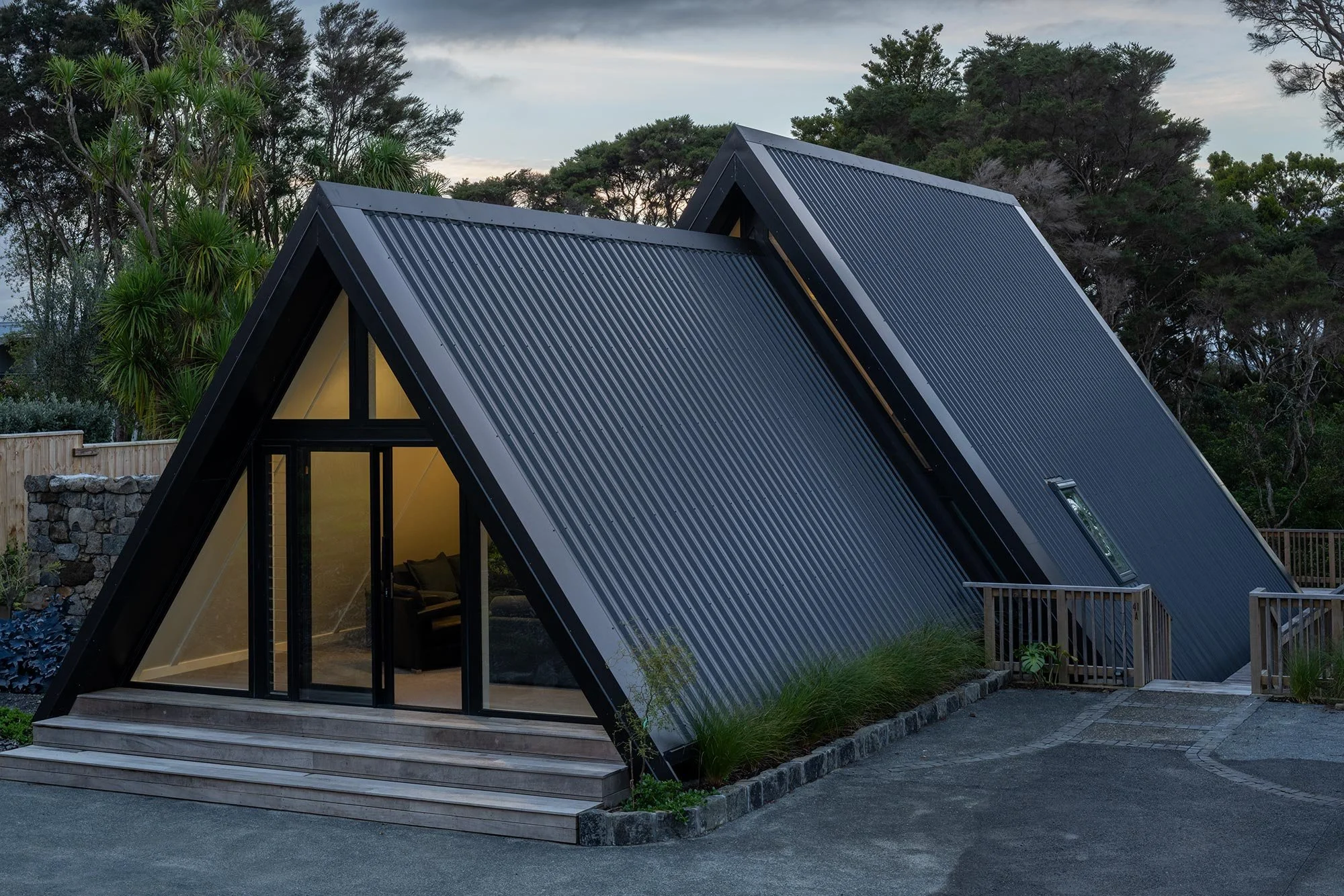 A modern new build A-frame house in Auckland with large glass doors and windows, black exterior, and a wooden deck with steps, set outdoors with trees and sky in background.