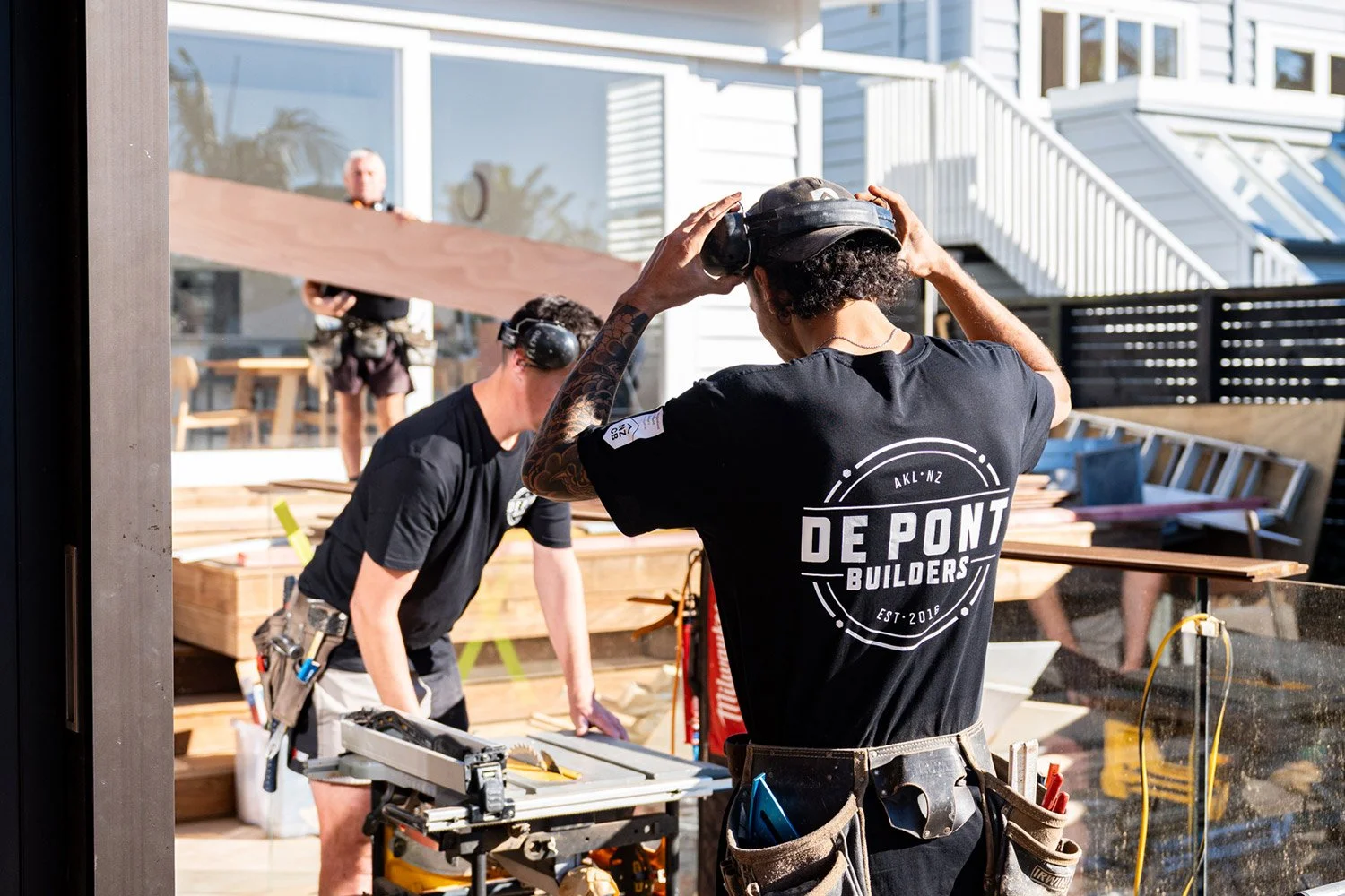 Two De Pont Builders construction workers building a house outdoors. One worker is adjusting their helmet, while another is working with tools on a workbench.