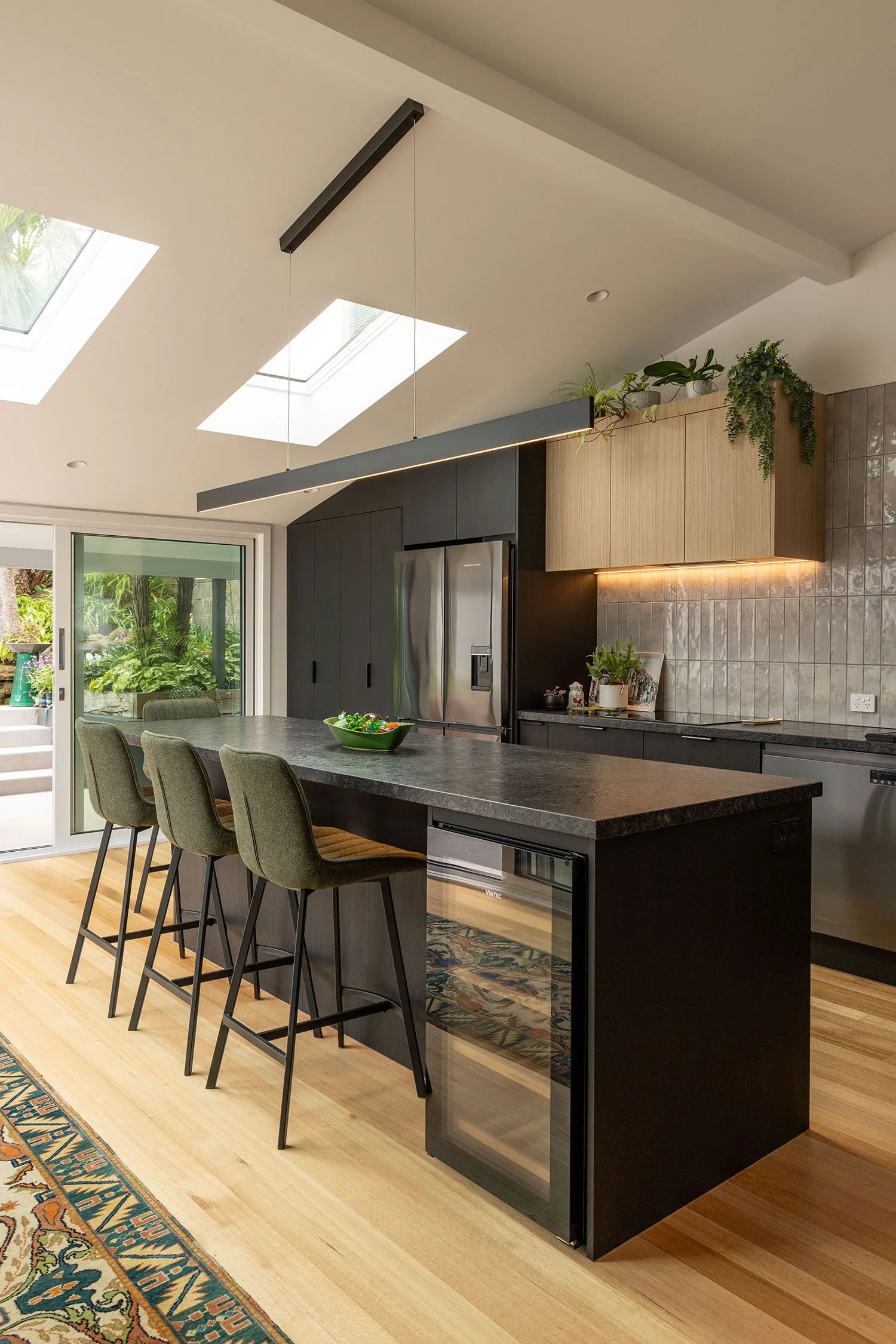 Modern renovated kitchen in Auckland, with black cabinets, stainless steel appliances, a concrete island with four gray bar stools, and a ceiling with skylights and a linear pendant light.
