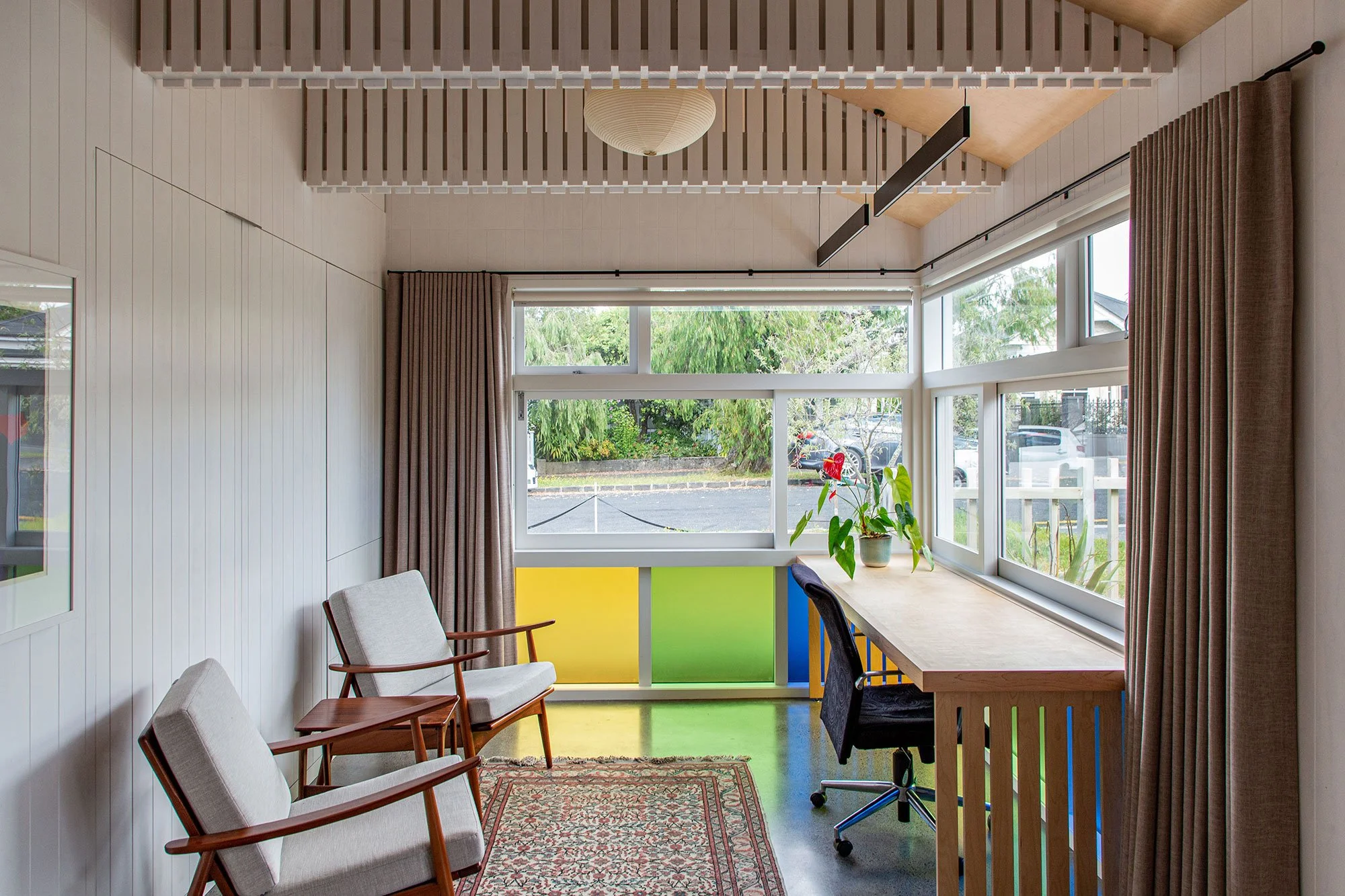 Interior view of a renovated multi-use room in Auckland with large windows, beige curtains, wooden chairs, a table, and a houseplant, with a colorful partition and ceiling light.