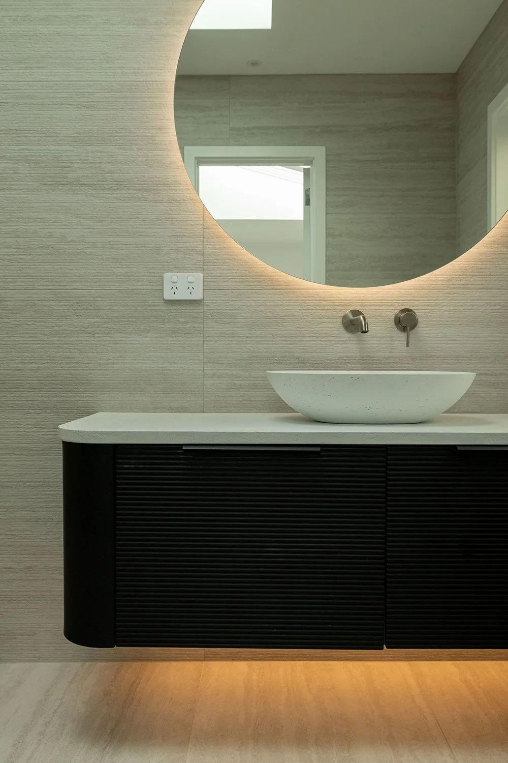 Modern renovated bathroom in Auckland with a round mirror, floating black vanity with ribbed doors, a countertop vessel sink, wall-mounted faucet, and a light-colored textured wall.