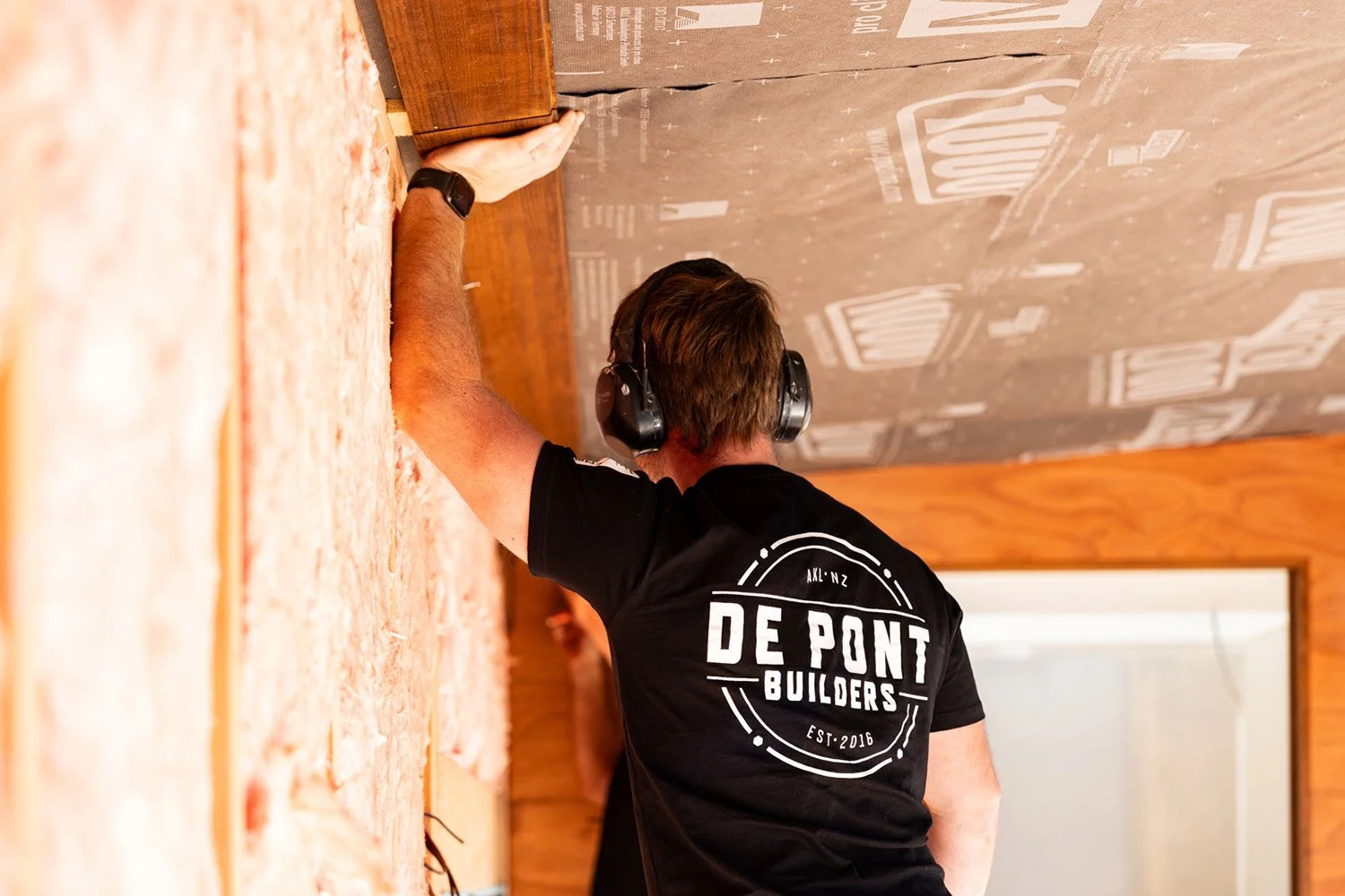 De Pont Builders' Construction worker wearing protective ear muffs and a black t-shirt with 'De Pont Builders' on the back, working on a renovation in Auckland.