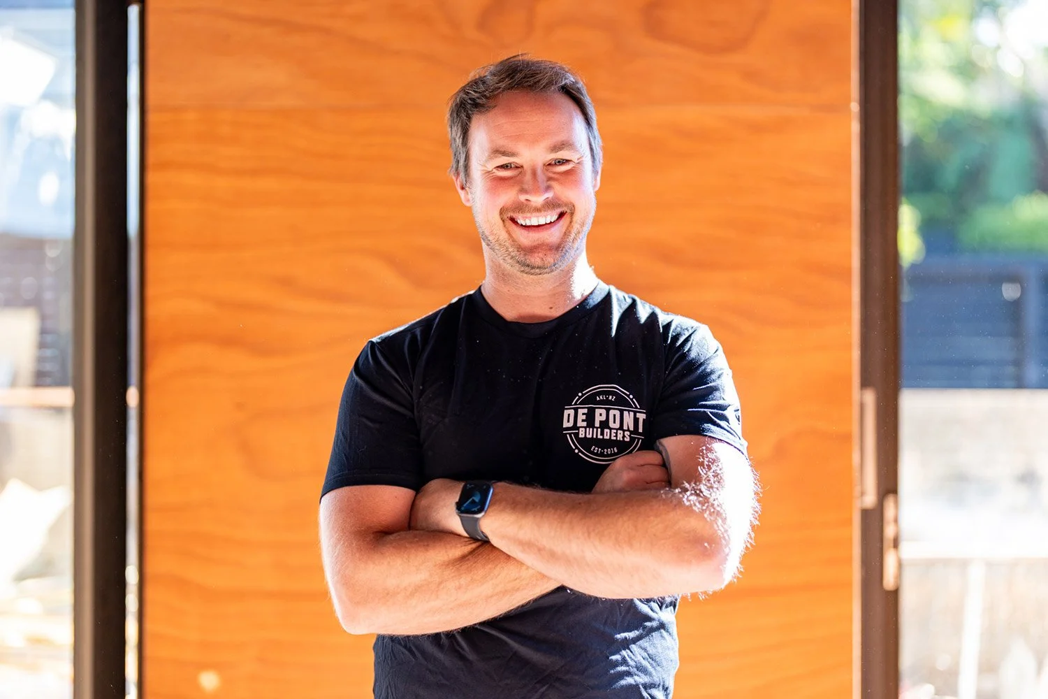 Josh De Pont, wearing a black T-shirt with a logo that says "De Pont Builders," standing in front of a wooden wall with glass doors on either side.
