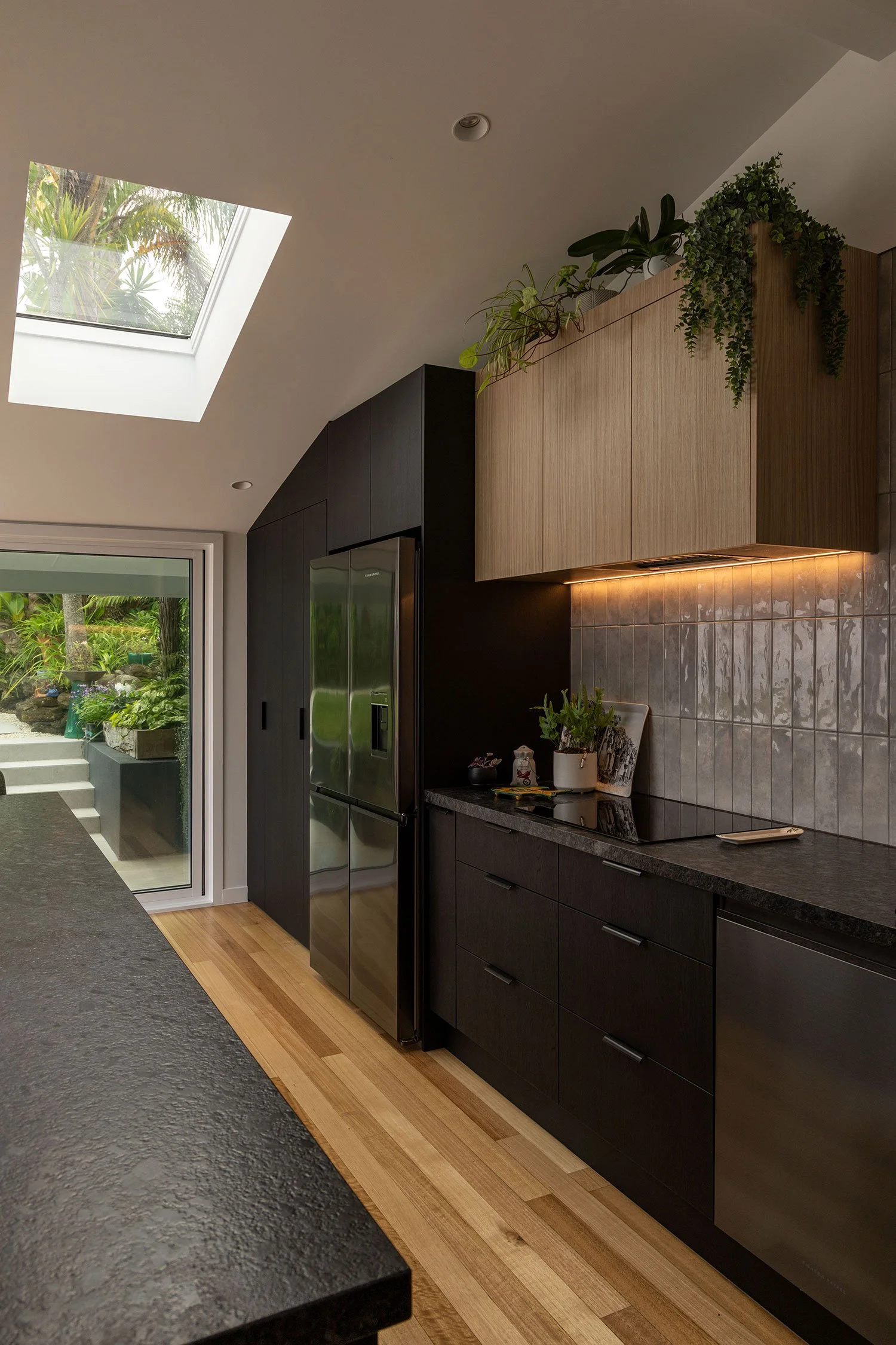 Modern renovated kitchen extension in Auckland with dark cabinets, wood upper cabinets, stainless steel refrigerator, tile backsplash, and houseplants on top of cabinets. Skylight and sliding glass door reveal outdoor garden scenery.