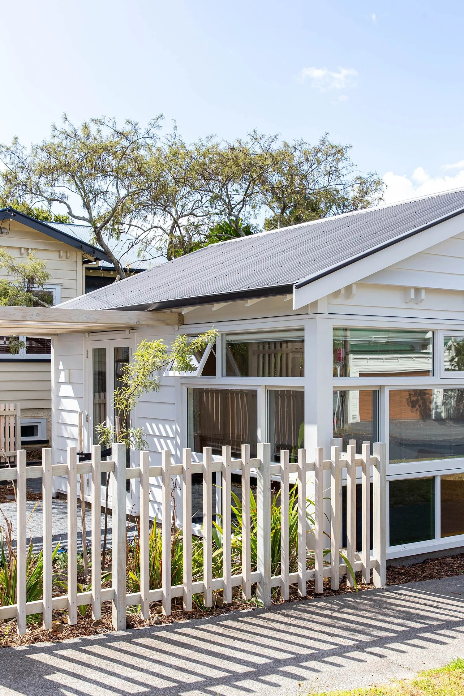 A charming house in Auckland with a white fence in front, surrounded by greenery and trees, under a bright blue sky.