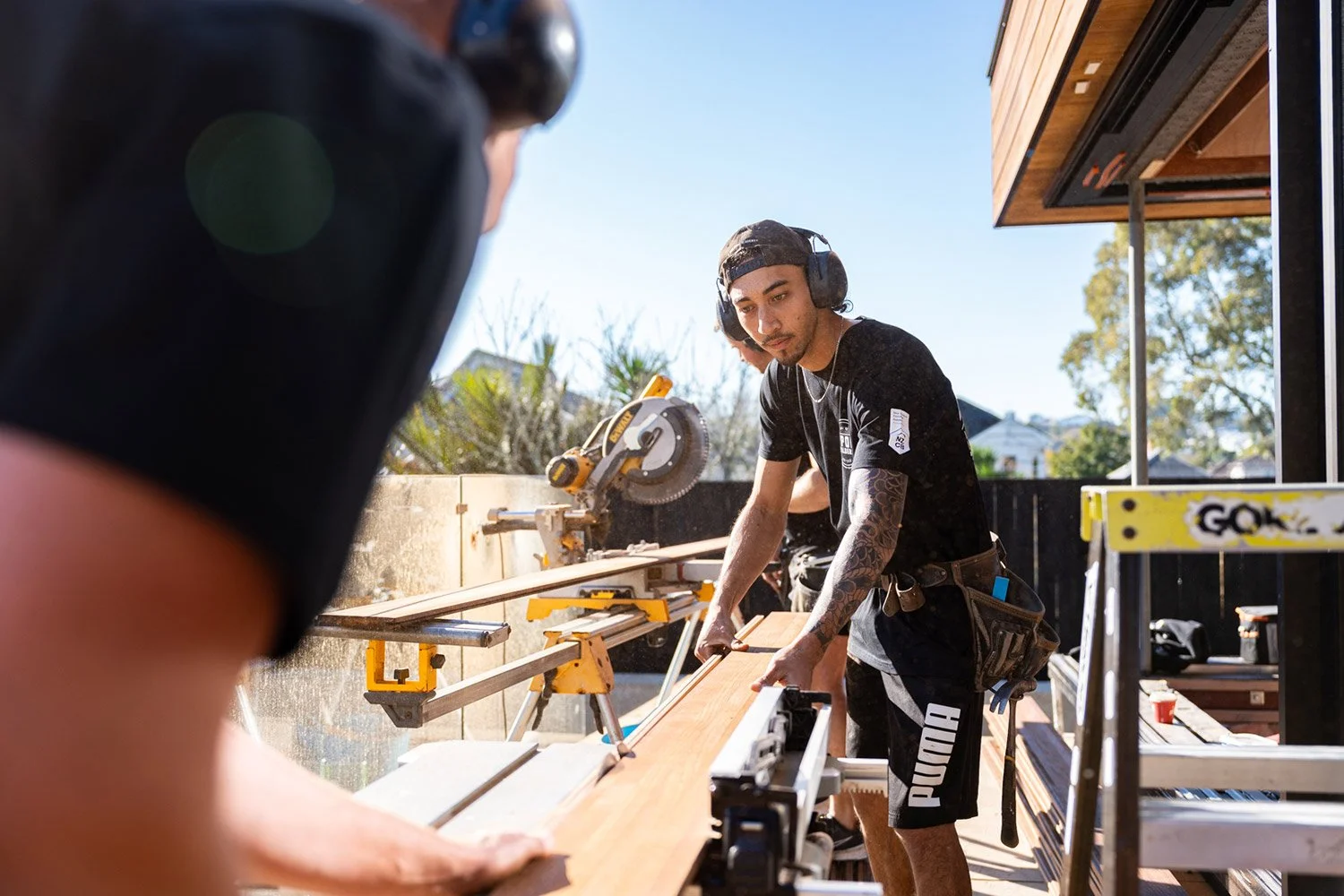 Two De Pont Builders construction workers. One is using a saw to cut a piece of timber on a worktable. The other is helping guide the timber.