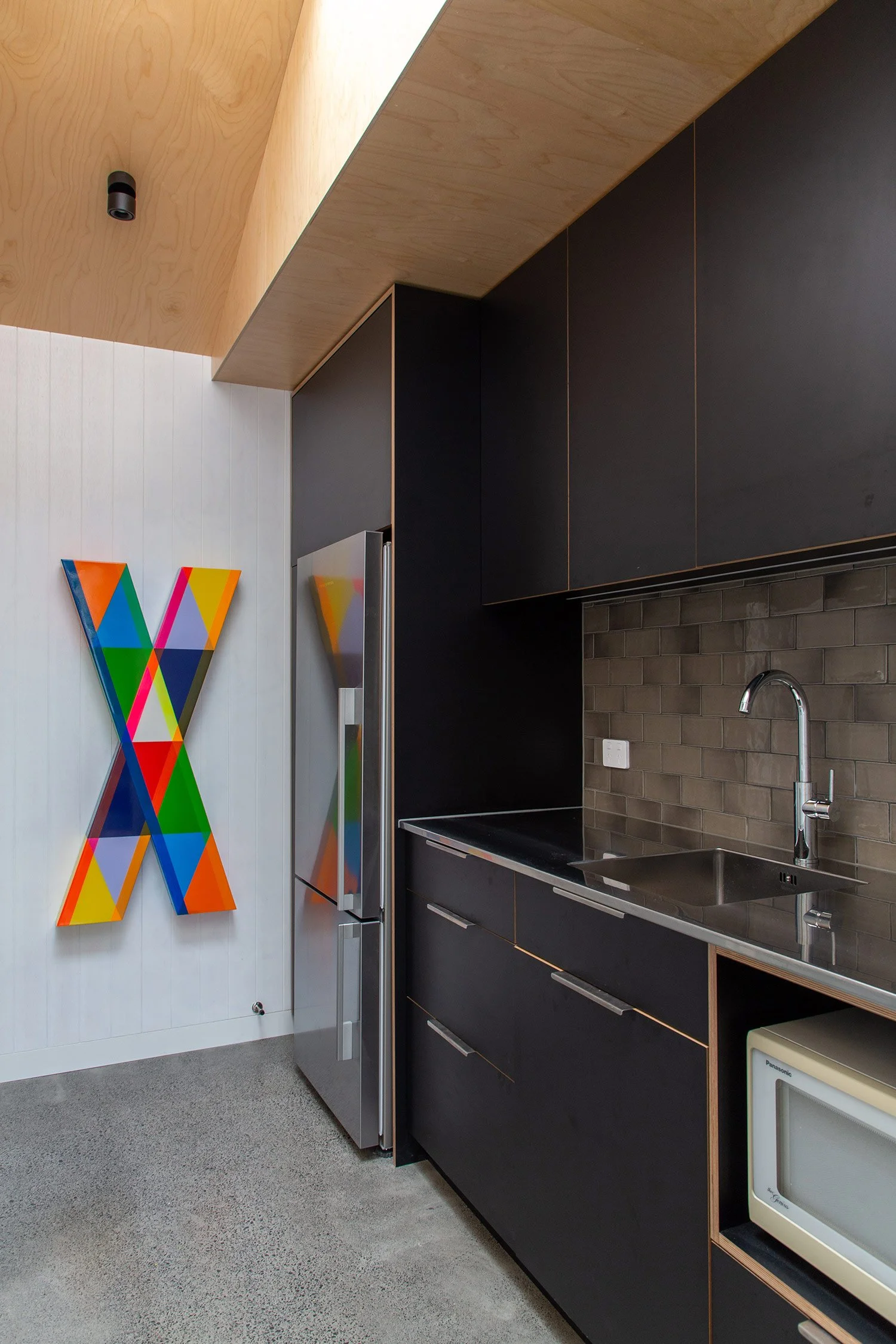Renovated kitchen in Auckland with black cabinets, a stainless steel refrigerator, a gray countertop, and a brown brick backsplash. Colorful geometric art decorates a white wall.