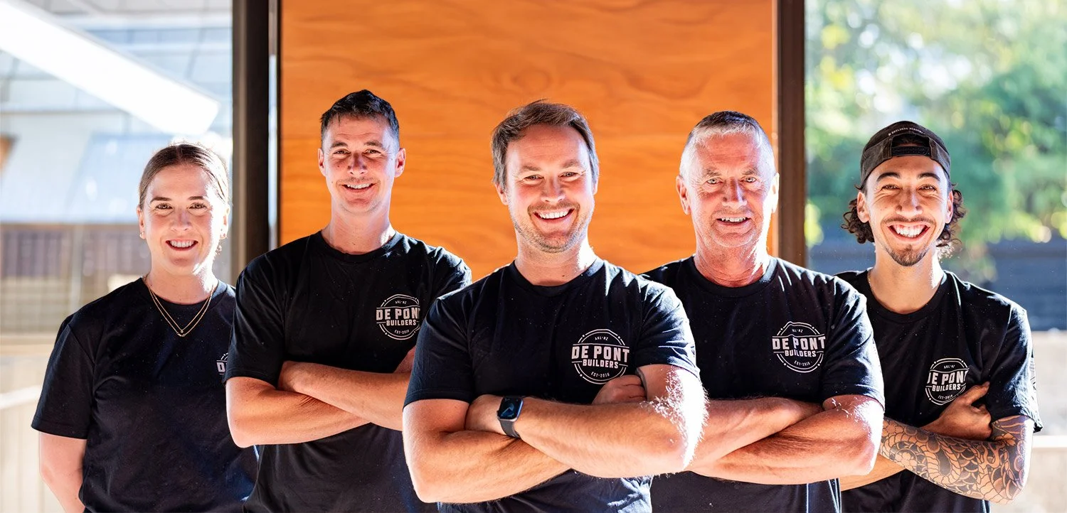 Group of five builders/De Pont Builders staff members standing indoors in front of a large window, wearing matching black t-shirts with the logo 'De Pont Builders'.