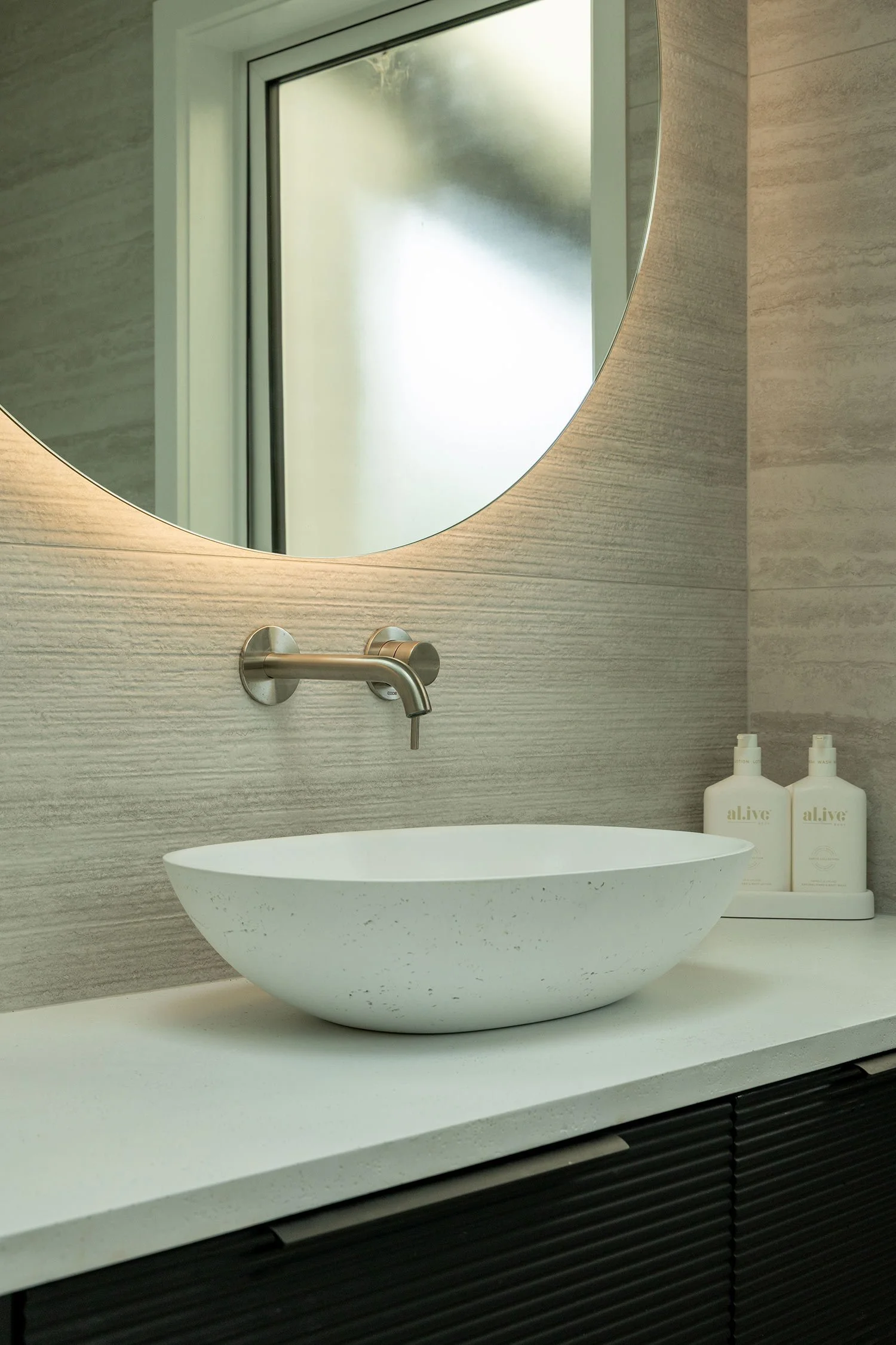 A modern bathroom renovation showing the sink area featuring a white, oval vessel sink on a white countertop, with a silver wall-mounted faucet above it. A round mirror is mounted on the wall.