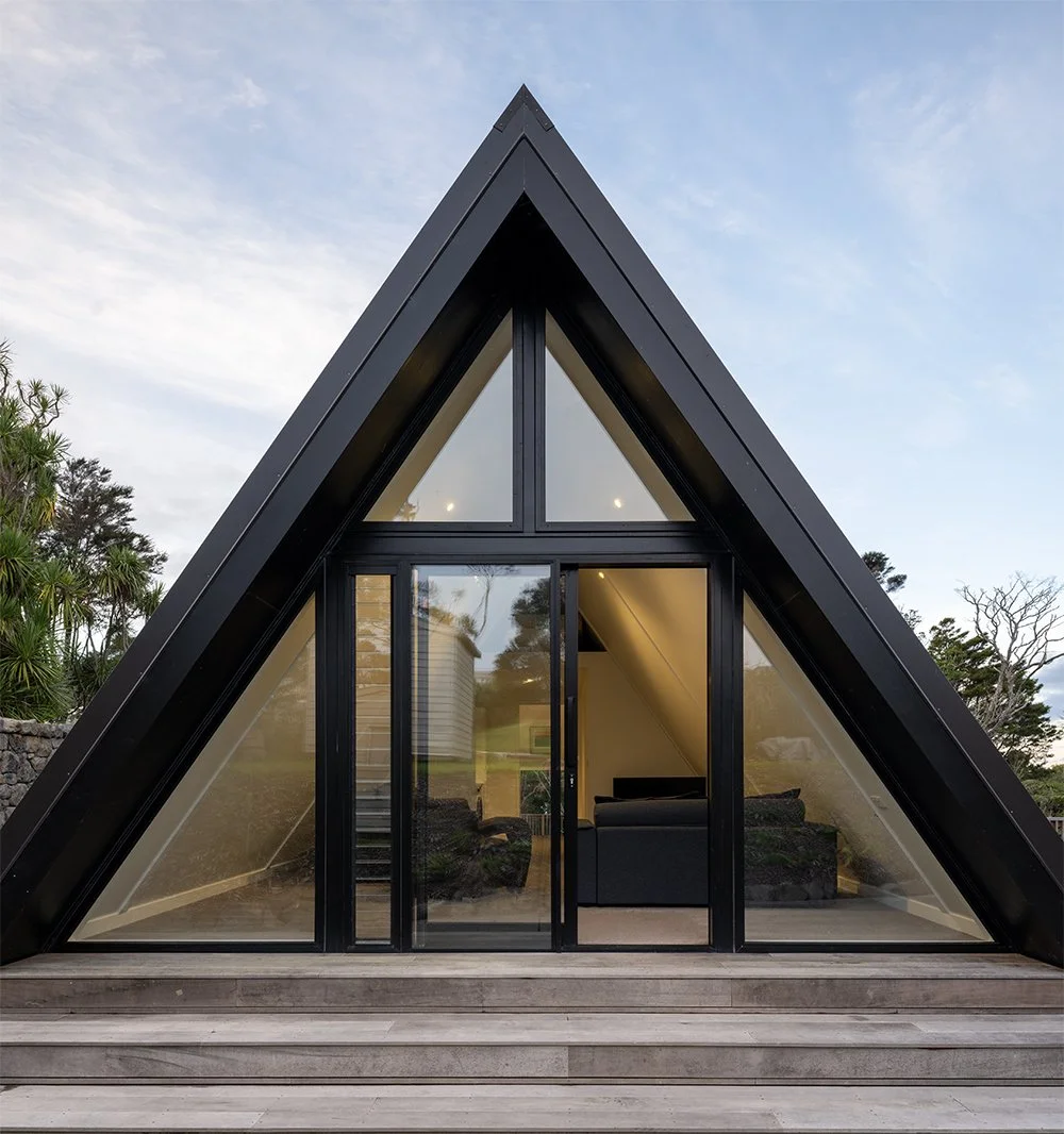 A modern new build A-frame house in Auckland with large glass doors and windows, black exterior, and a wooden deck with steps, set outdoors with trees and sky in background.