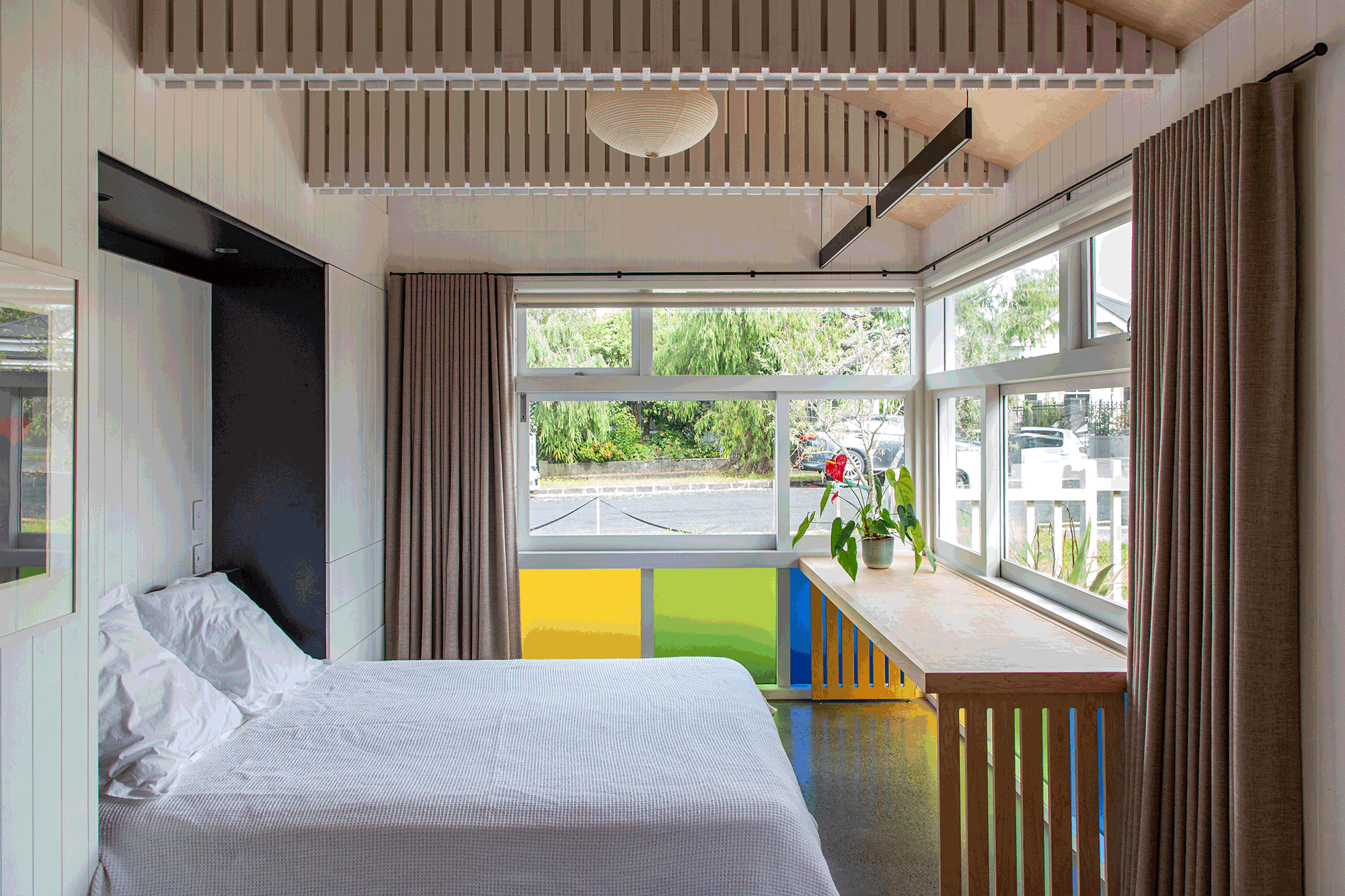 Interior view of a renovated multi-use room in Auckland with large windows, beige curtains, wooden chairs, a table, and a houseplant, with a colorful partition and ceiling light.