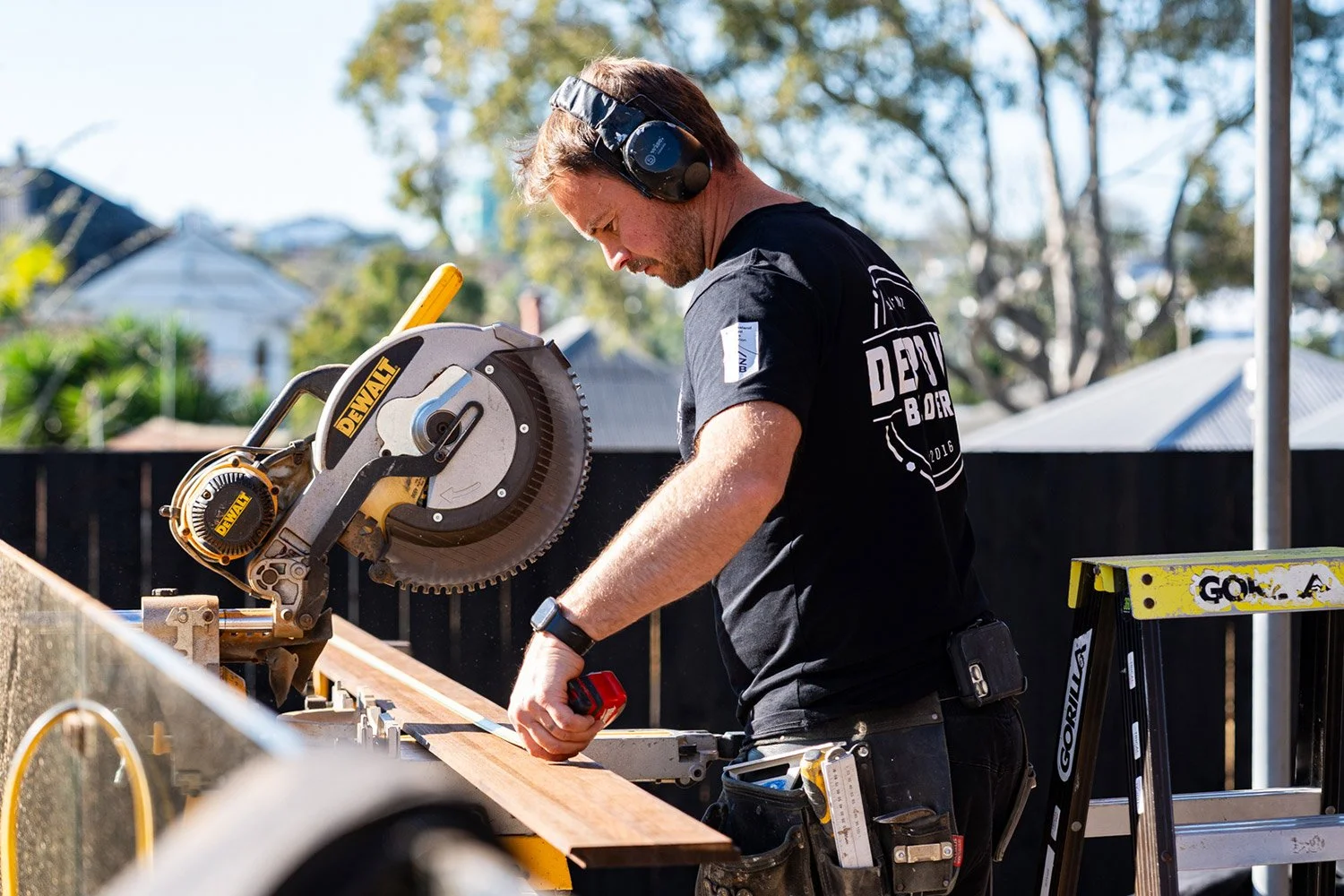 Josh De Pont working on an Auckland renovation, wearing ear protection, measuring a piece of timber with tools attached to his belt and a ladder nearby.