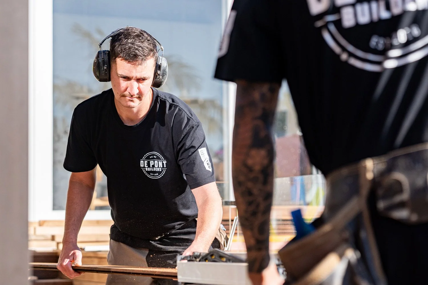 Two De Pont Builders construction workers. One wearing noise-canceling headphones and a black T-shirt with De Pont Builders logo, working on an Auckland renovation, with another person partially visible in the foreground.