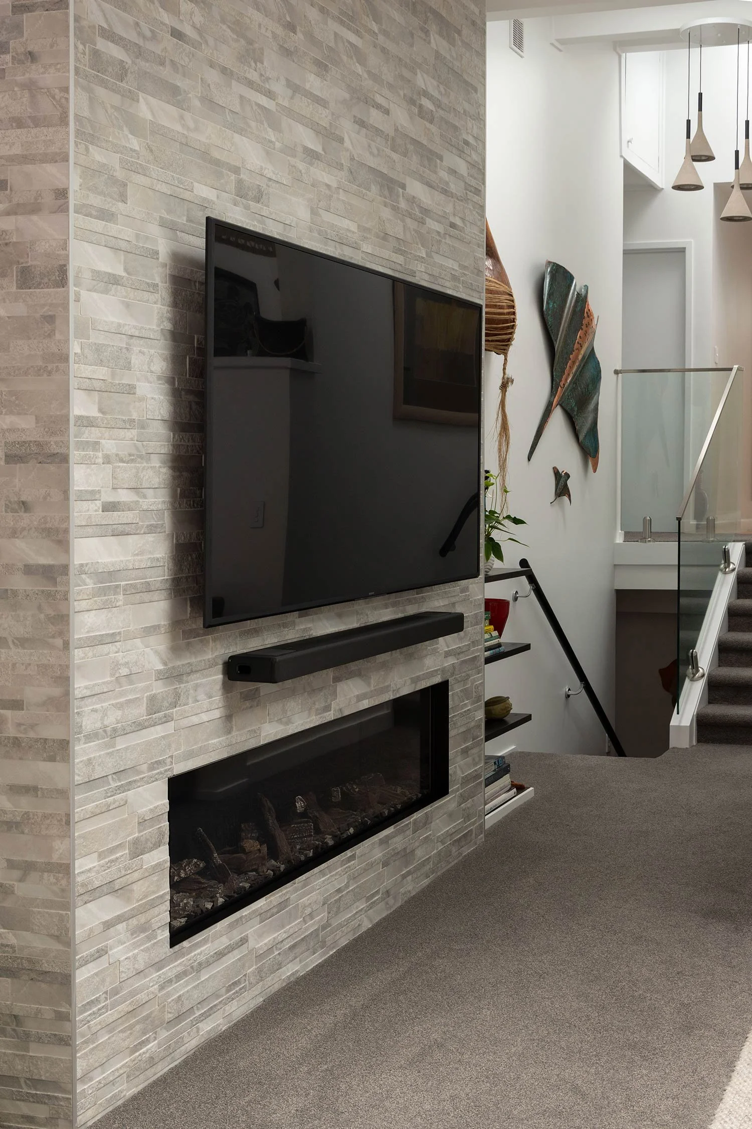 Interior of a modern recently renovated home in Auckland showing a stone fireplace, a wall-mounted television, and decorative artwork on the wall near a staircase with glass railing.