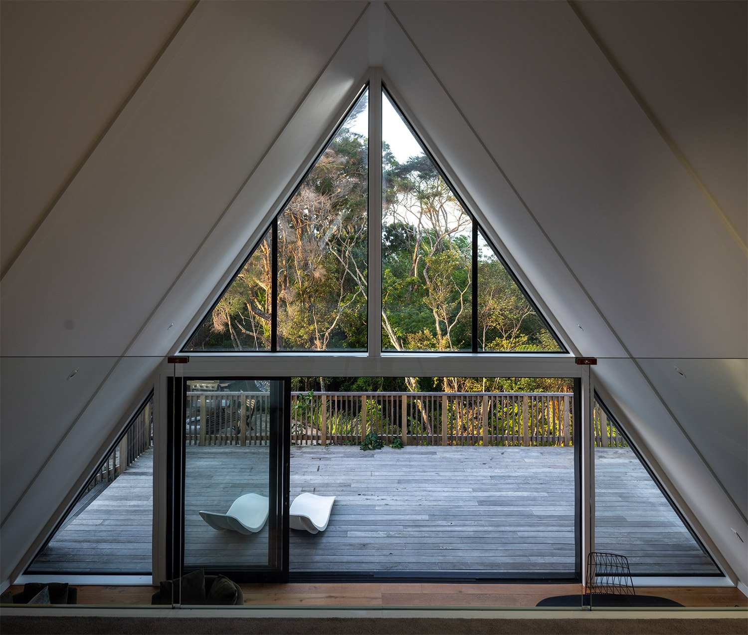 View from an interior room of a new build home in Auckland with a large triangular window, overlooking a wooden deck with two white lounge chairs and a forested area with trees.