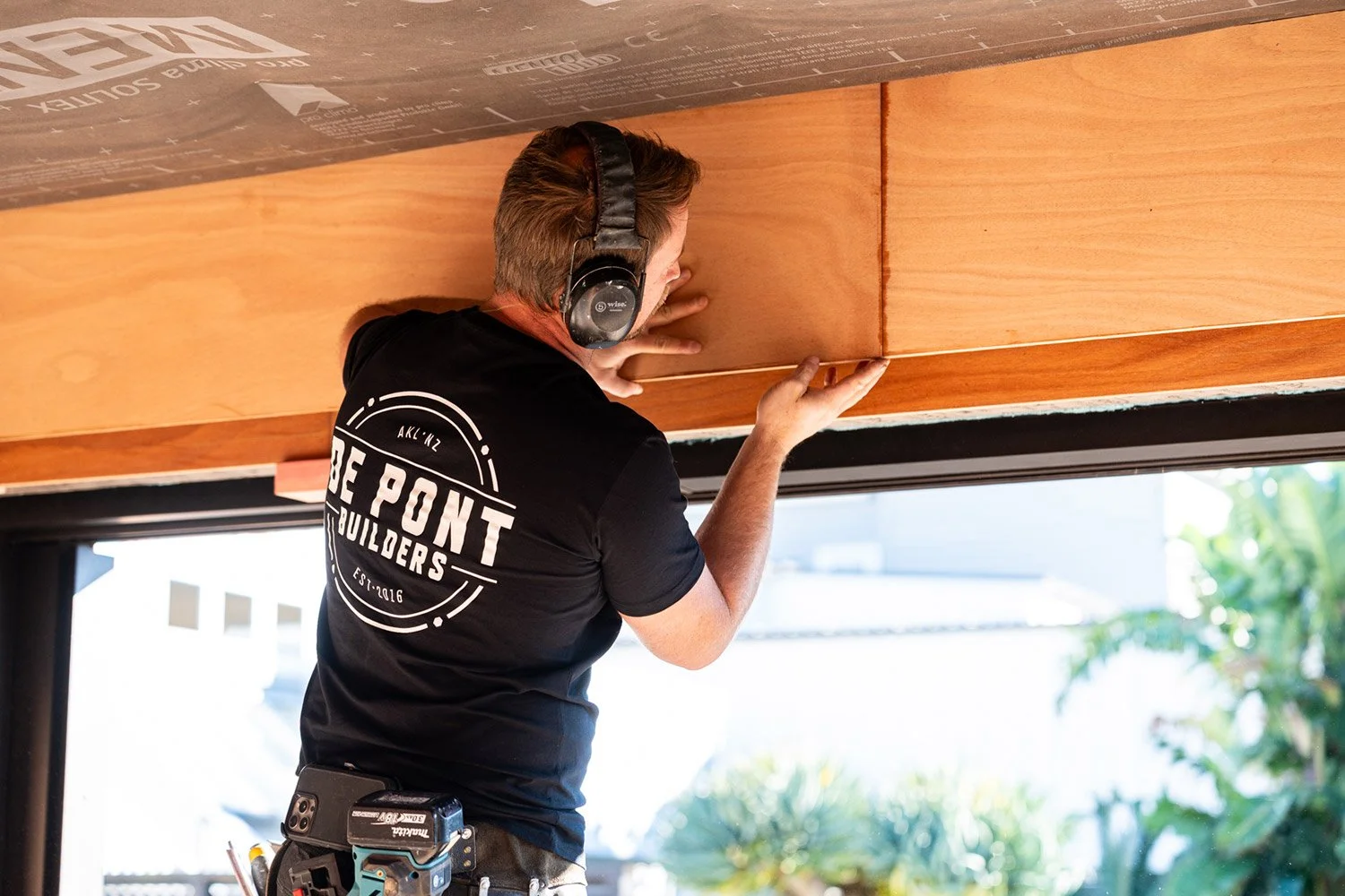 De Pont Builders' Construction worker installing wooden paneling, wearing headphones and a black T-shirt with 'The Point Builders' logo, tools attached to belt, in a bright indoor space in Auckland with large window showing outdoor greenery.