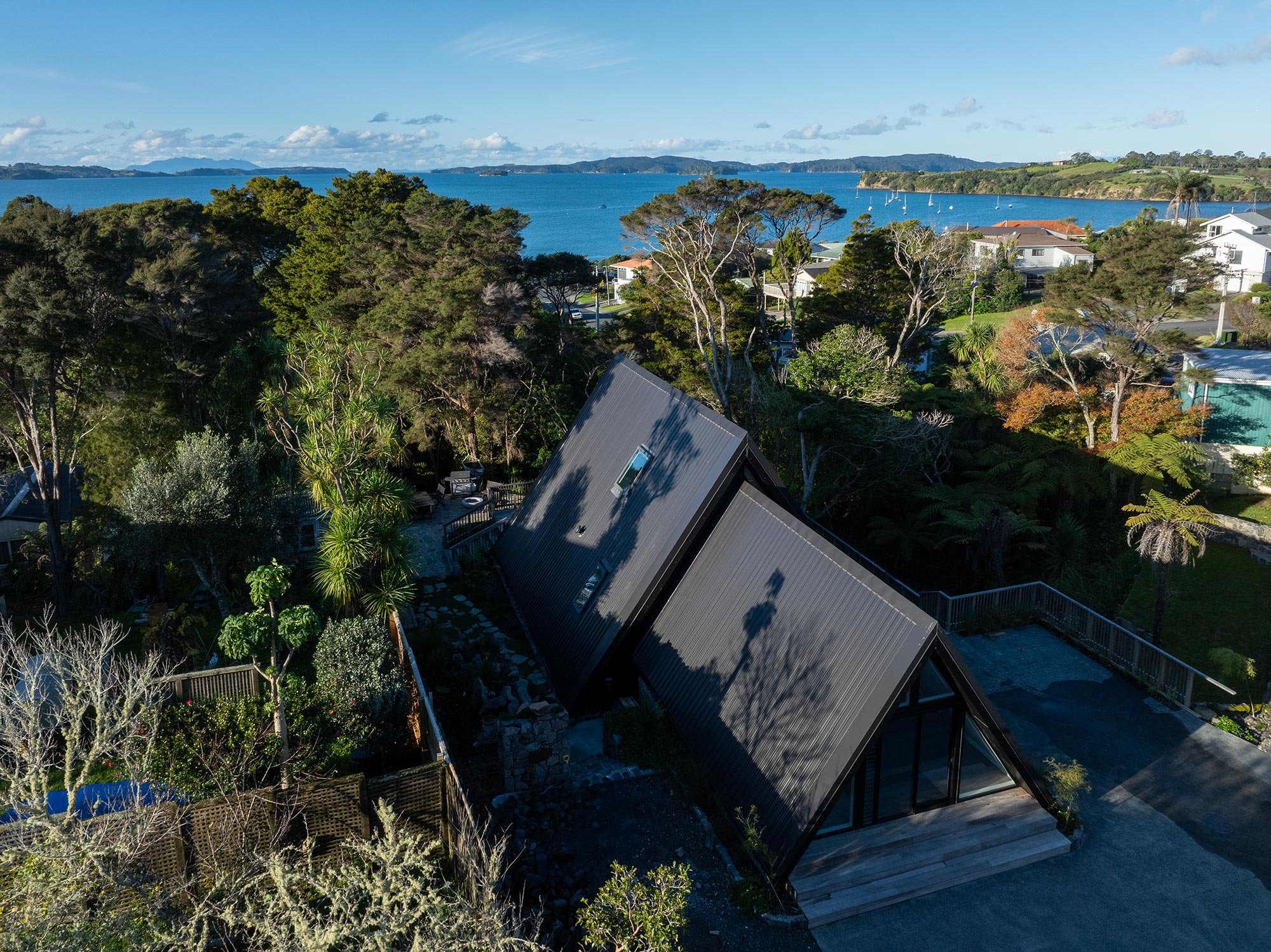 A modern new build A-frame house in Auckland with black exterior. There are sailboats on the water and hills in the distance under a partly cloudy sky.