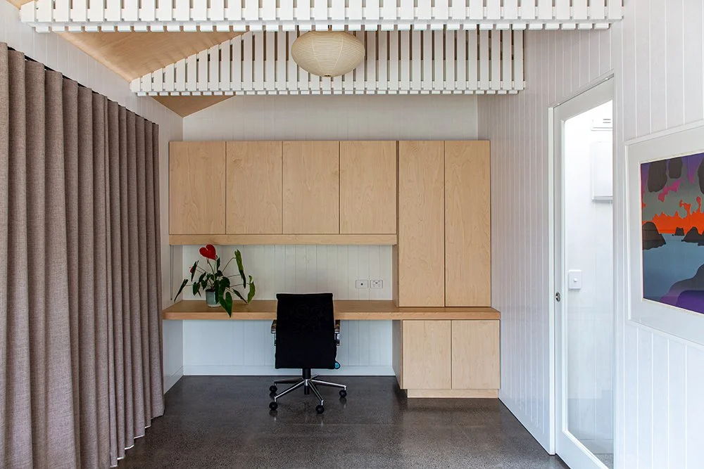A renovated Auckland home office with modern built-in wooden cabinets, a desk, a black swivel chair, a potted plant with red flowers and beige curtains.