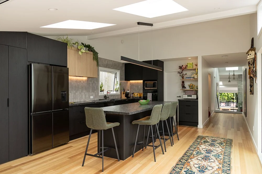 Modern renovated kitchen in Auckland with black and wood cabinets, a black refrigerator, gray countertops, and a kitchen island with four chairs. Includes skylights and decorative wall art.