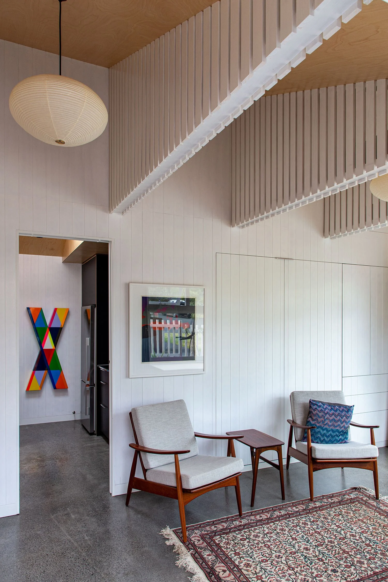Renovated multi-use space in Auckland with two mid-century modern style armchairs, a patterned rug, and a wall with framed art and architectural slat design ceiling.