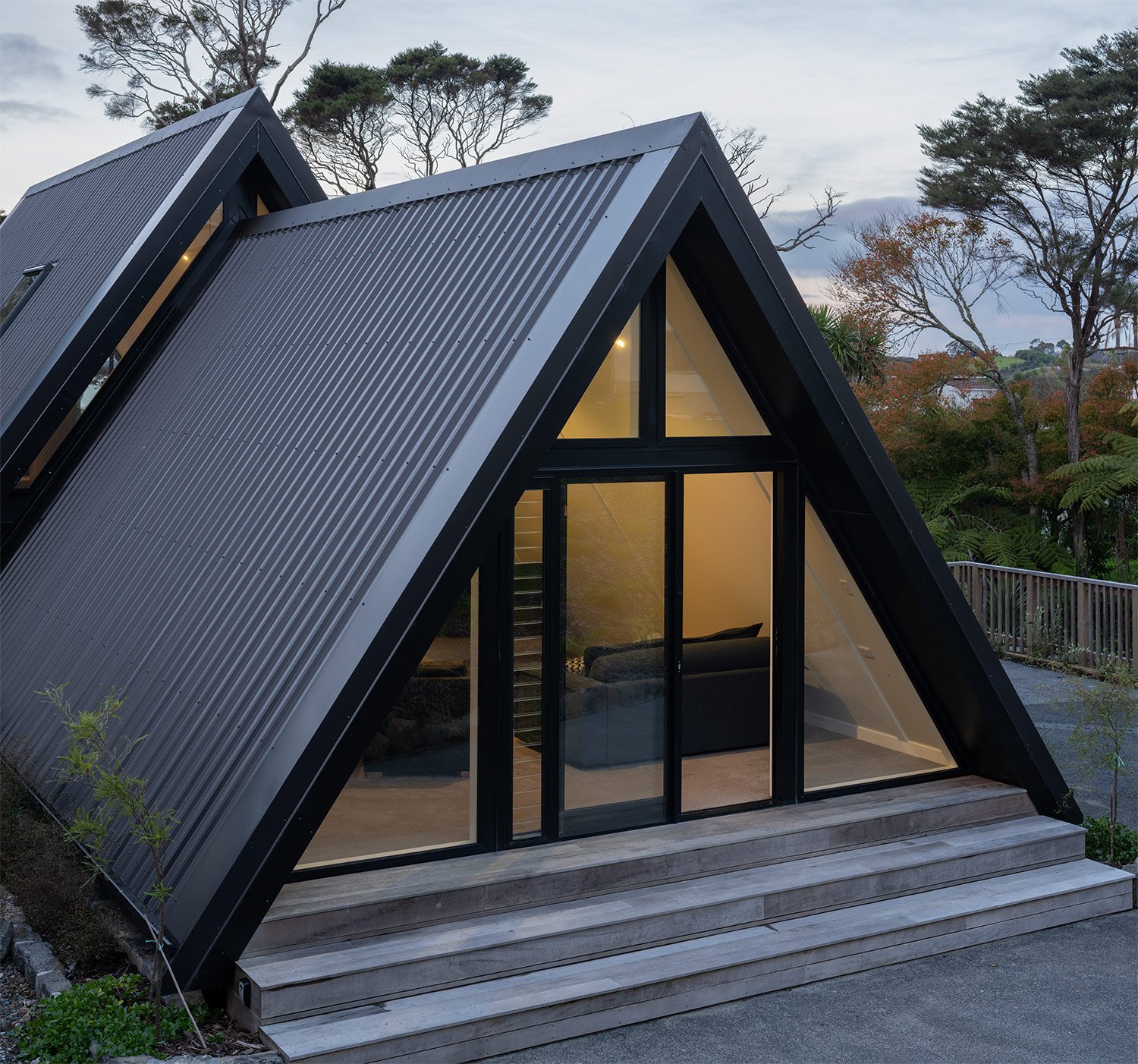 A modern new build A-frame house in Auckland with large glass doors and windows, black exterior, and a wooden deck with steps, set outdoors with trees and sky in background.