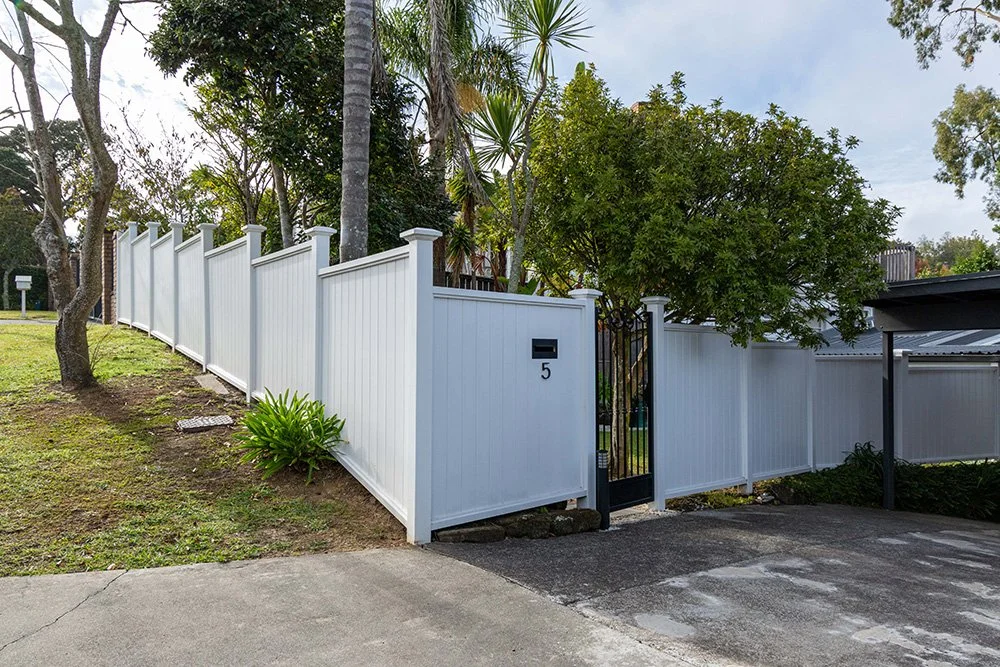 A white modern and well built privacy fence in Auckland surrounding a yard. Trees and greenery nearby, concrete driveway.