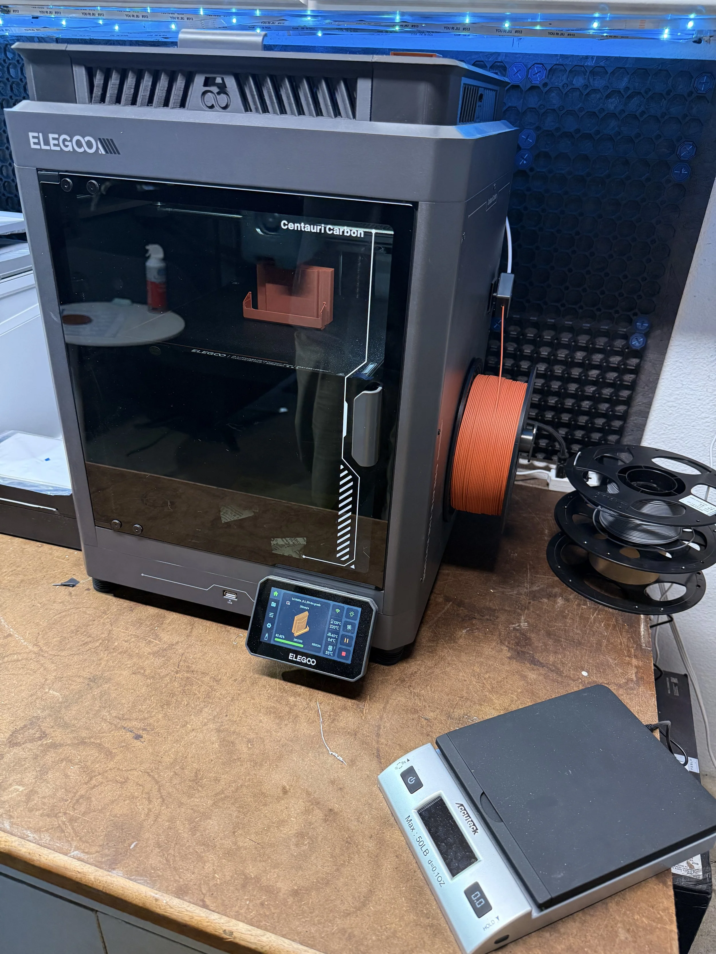 3D printer in a workshop, with spools of filament on the side, a touchscreen control panel displaying print details, and a box of filament on the desk.