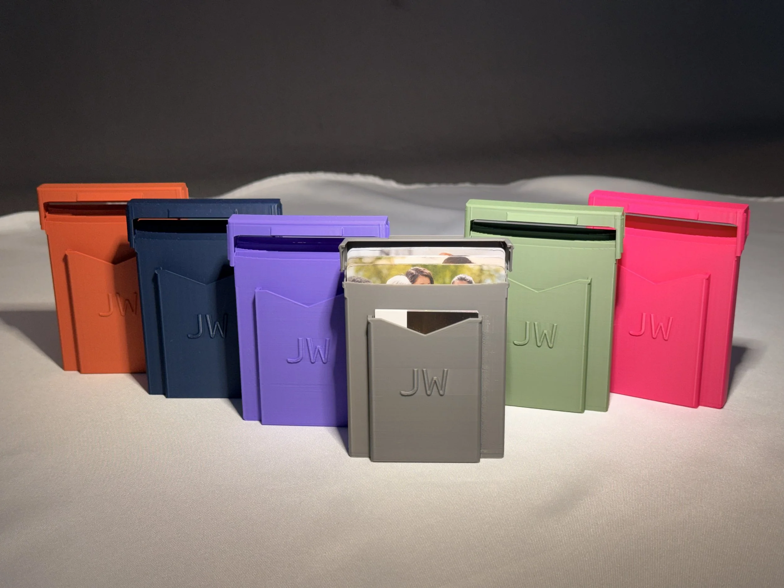 Colorful 3D printed JW tract holders in orange, blue, purple, gray, green, and pink arranged on a gray surface with a dark background.