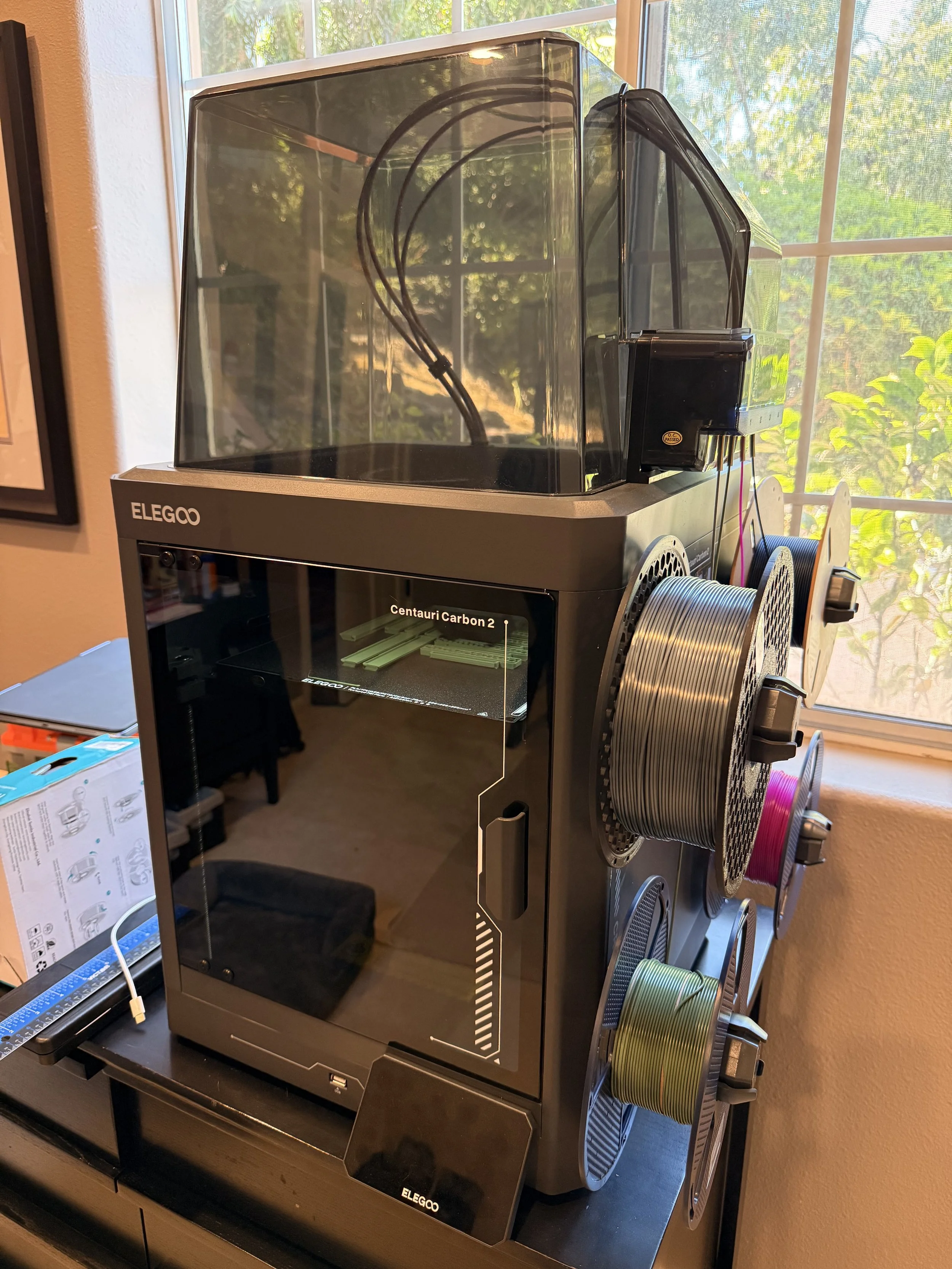 A 3D printer with spools of filament on its side, situated near a window with natural light illuminating the area.