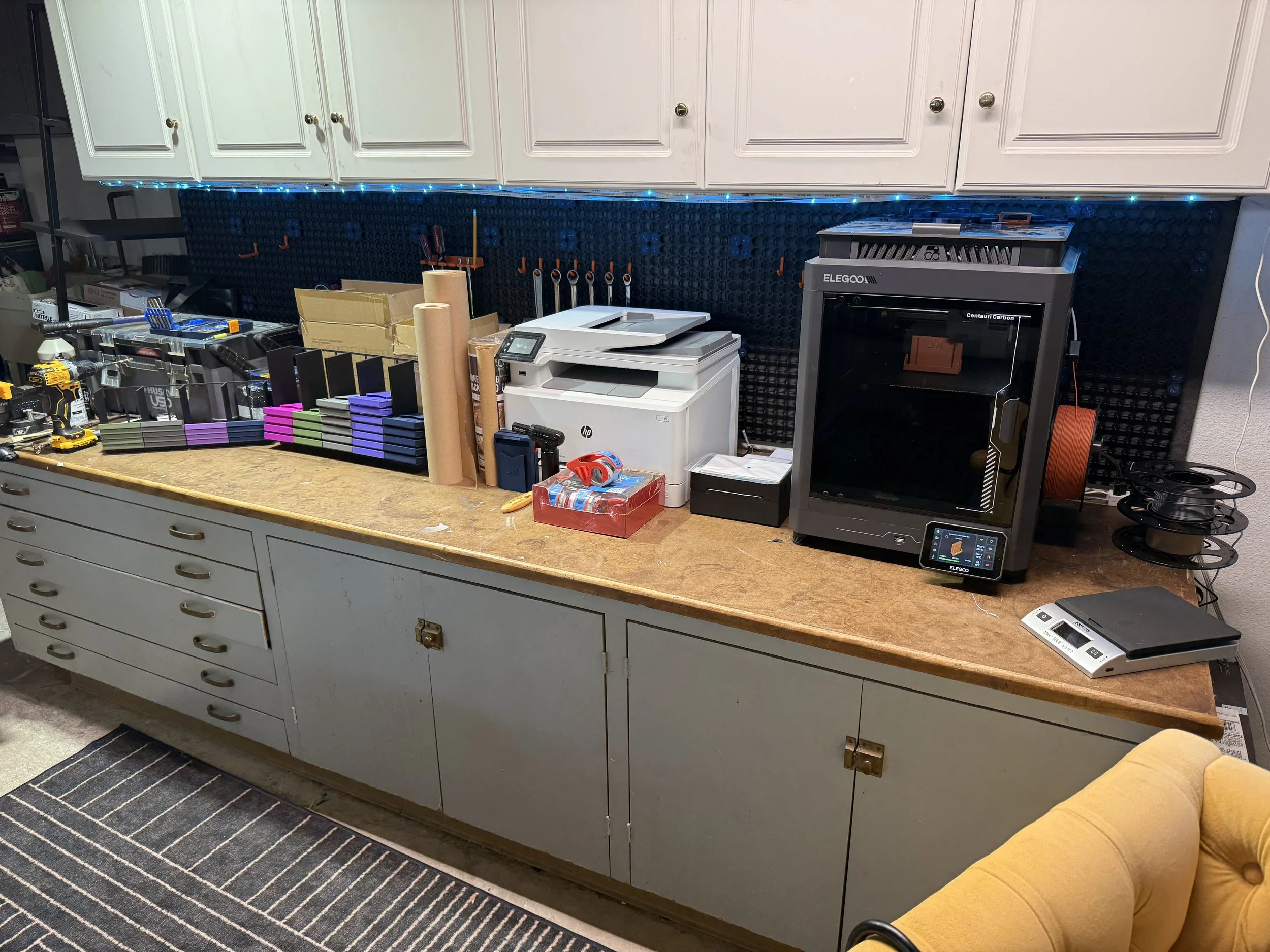 Workbenchtop with tools and supplies, including a printer, boxes, rolls of paper, screwdrivers, and a 3D printer, against a black pegboard with blue LED strip lighting. Cabinets and a yellow chair are also visible.