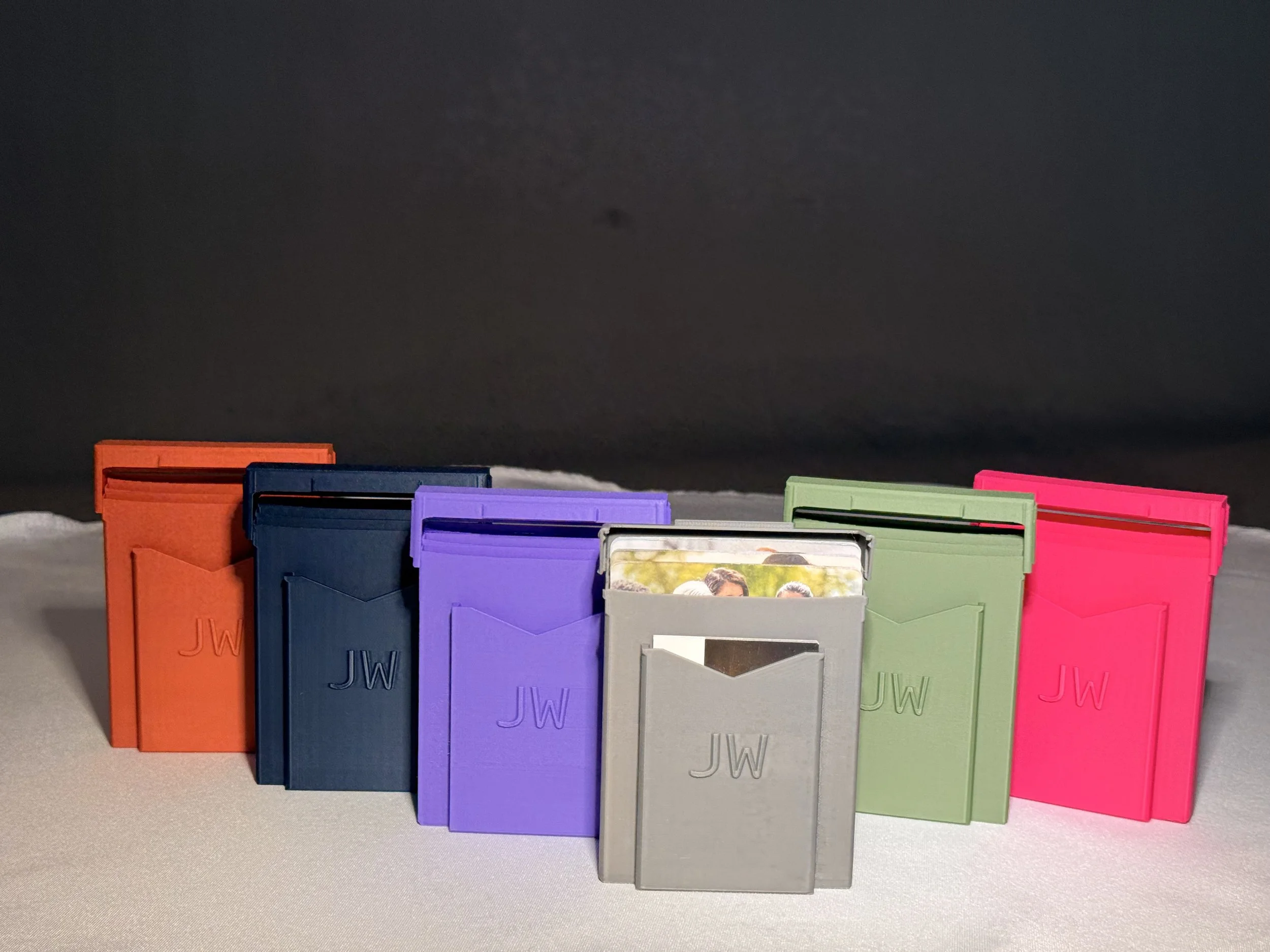 Colored tract holders arranged in a row on a surface, with a dark background. Each tract holder has the initials 'JW' embossed on the cover.