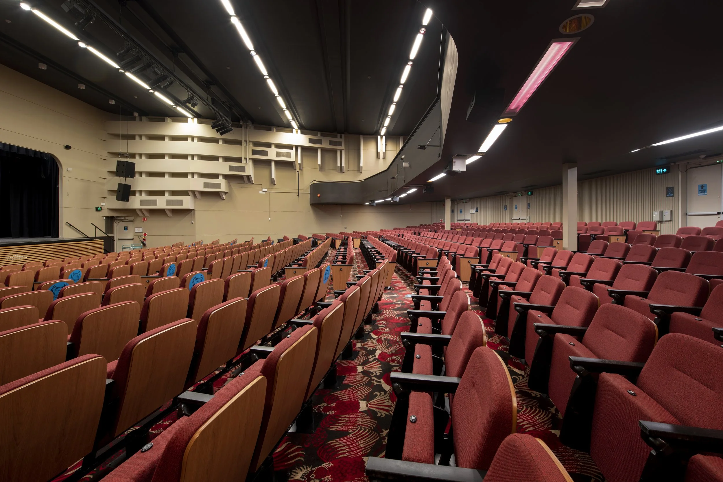 Interior of a large auditorium or theater with multiple rows of red cushioned seats, a stage on the left, and a high ceiling with lighting fixtures.