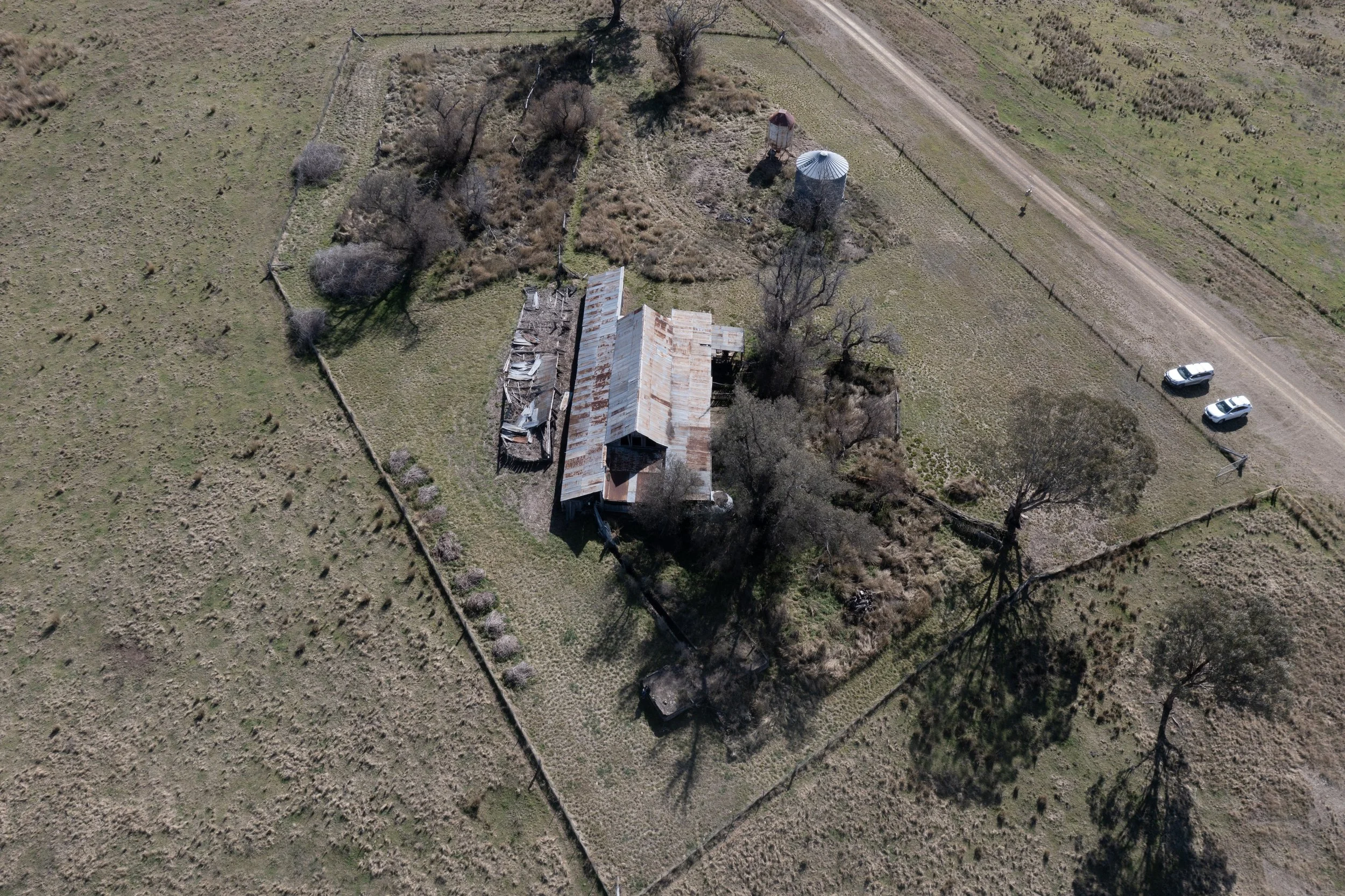 An aerial view of an old, rusty barn with a metal roof, surrounded by fenced grassland, with a dirt road and a few parked cars nearby. There are trees and small structures around the barn.