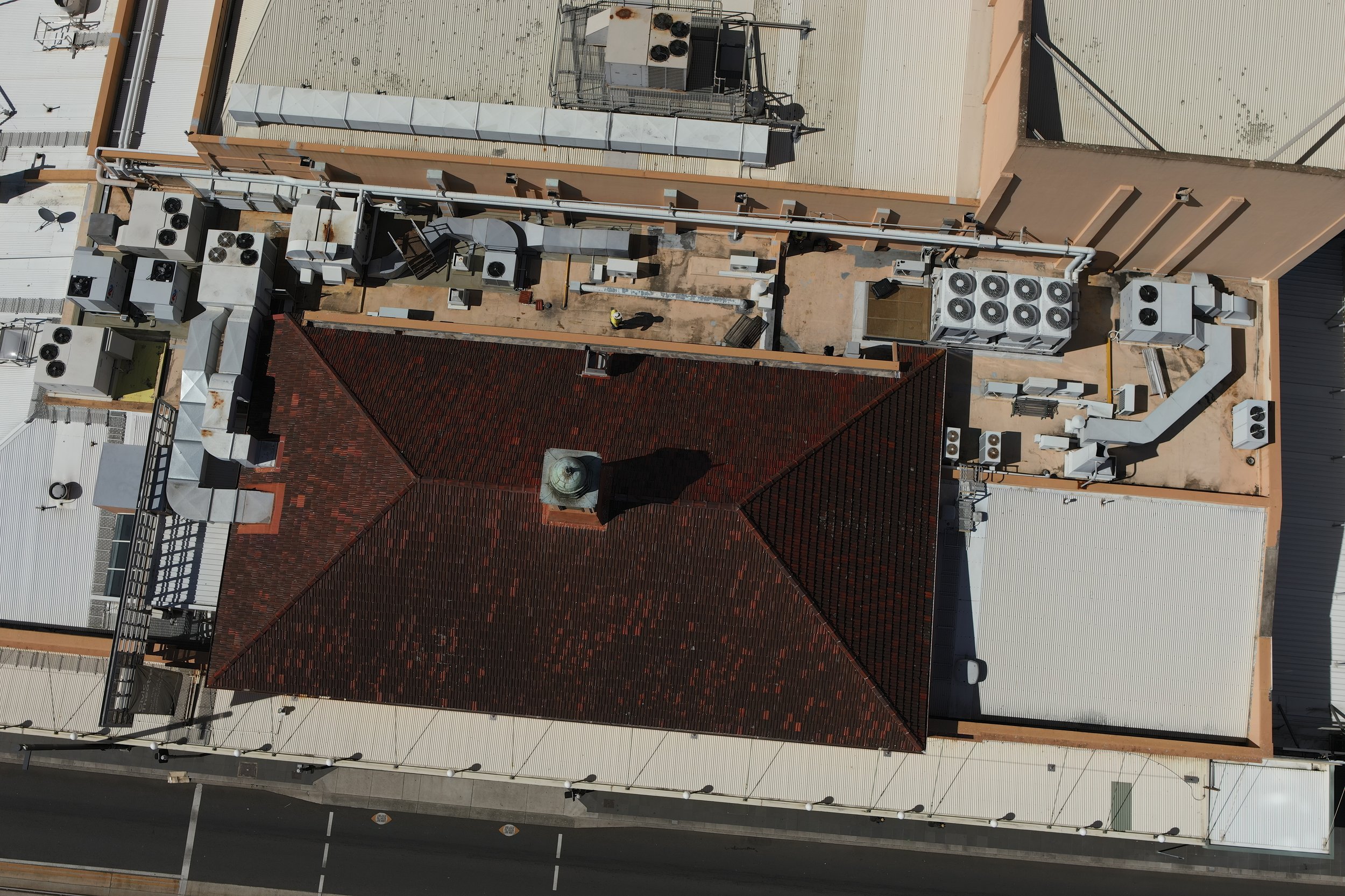 An aerial view of a rooftop with various HVAC units, ducts, and ventilation systems, adjacent to a red brick building with a water tower on top, and a street with parking spaces below.