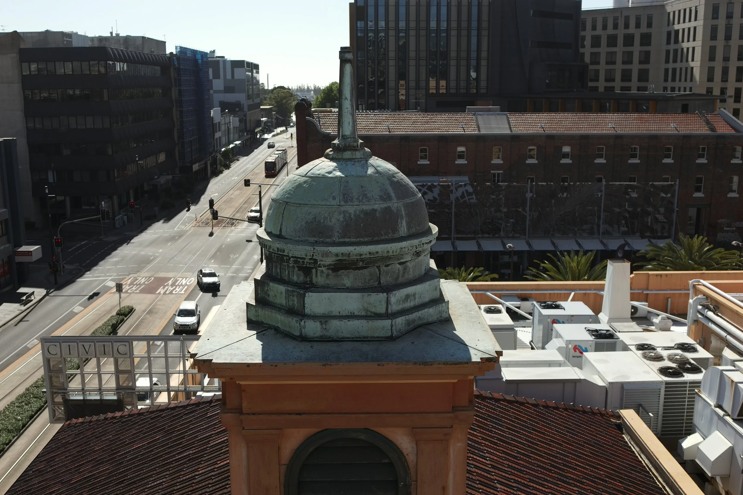 City street view seen from a rooftop, with a weathered decorative chimney in the foreground, surrounding modern buildings, traffic lights, cars, and a tram on the street.