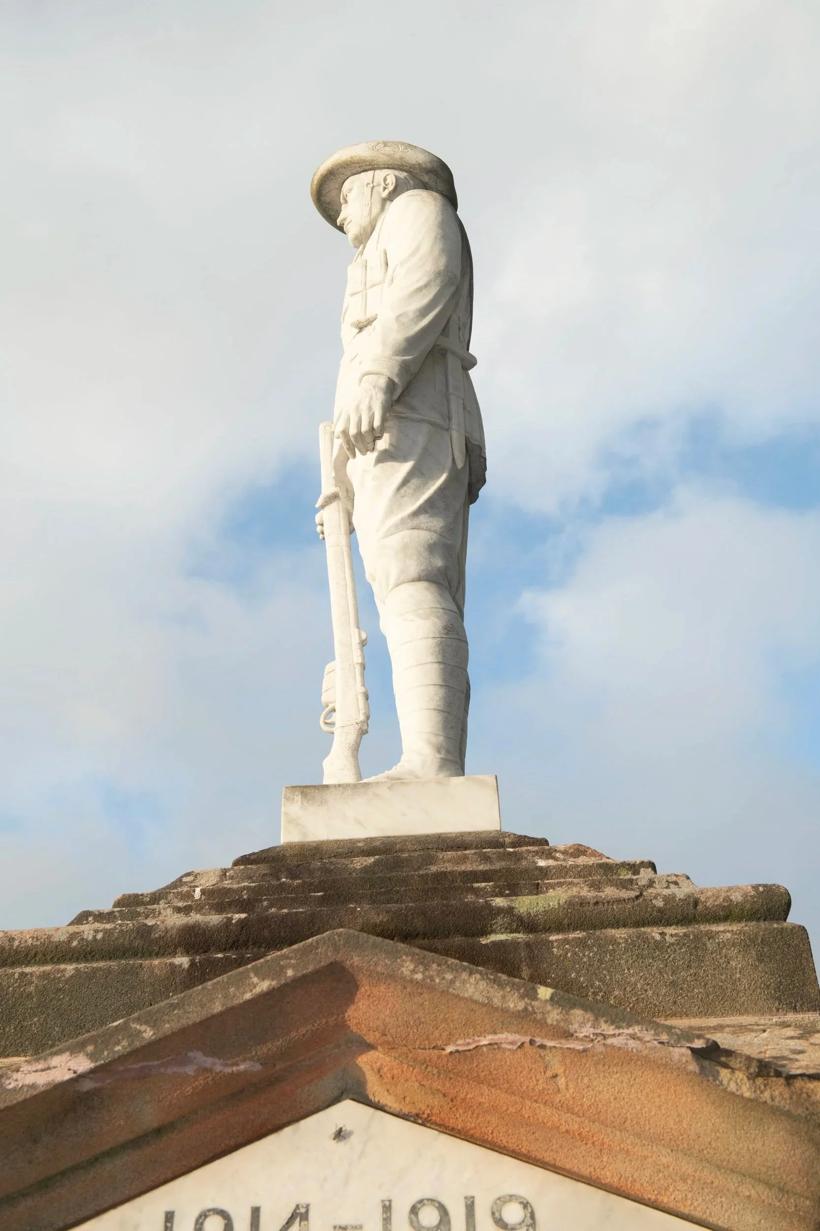 A tall white marble statue of a soldier holding a rifle, wearing a wide-brimmed hat, standing on a stone pedestal against a cloudy sky.