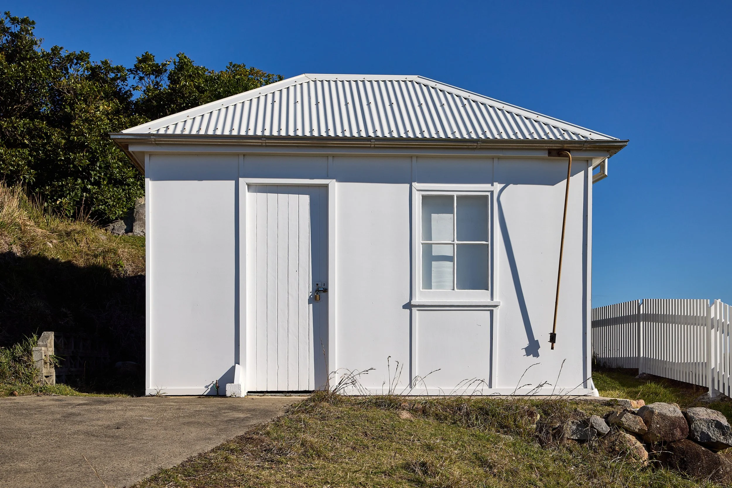 White small building with a corrugated metal roof, a single window, and a door, surrounded by a white picket fence and greenery, under a clear blue sky.