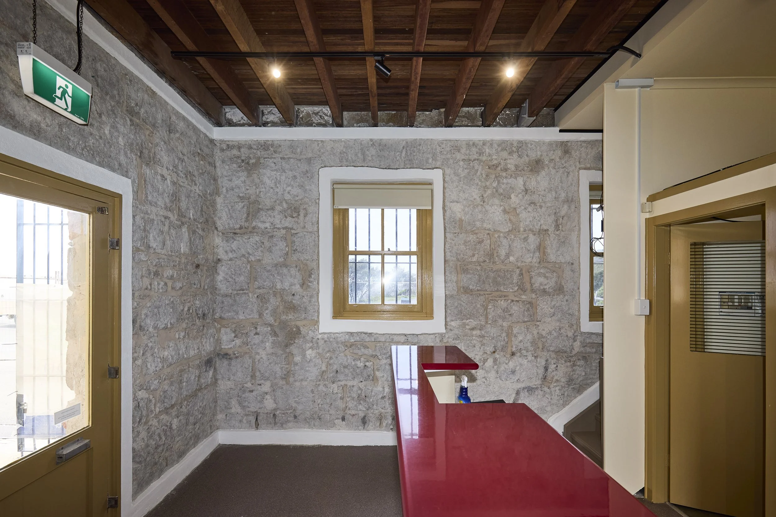 A room with exposed stone walls, a wooden ceiling with visible beams, two windows with beige frames, and a red countertop or table. There is an exit sign on the wall and some electrical wiring visible.