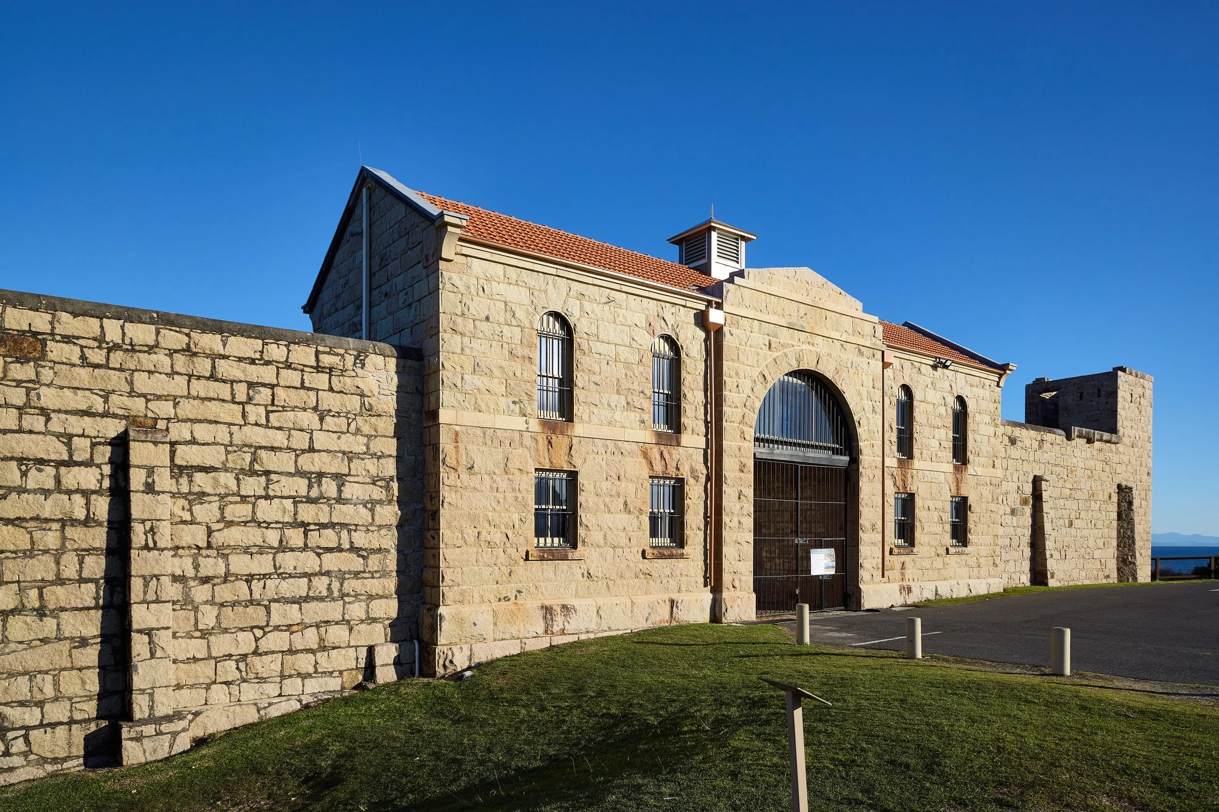 Stone building with arched windows, a gated entrance, and a small chimney under a clear blue sky.