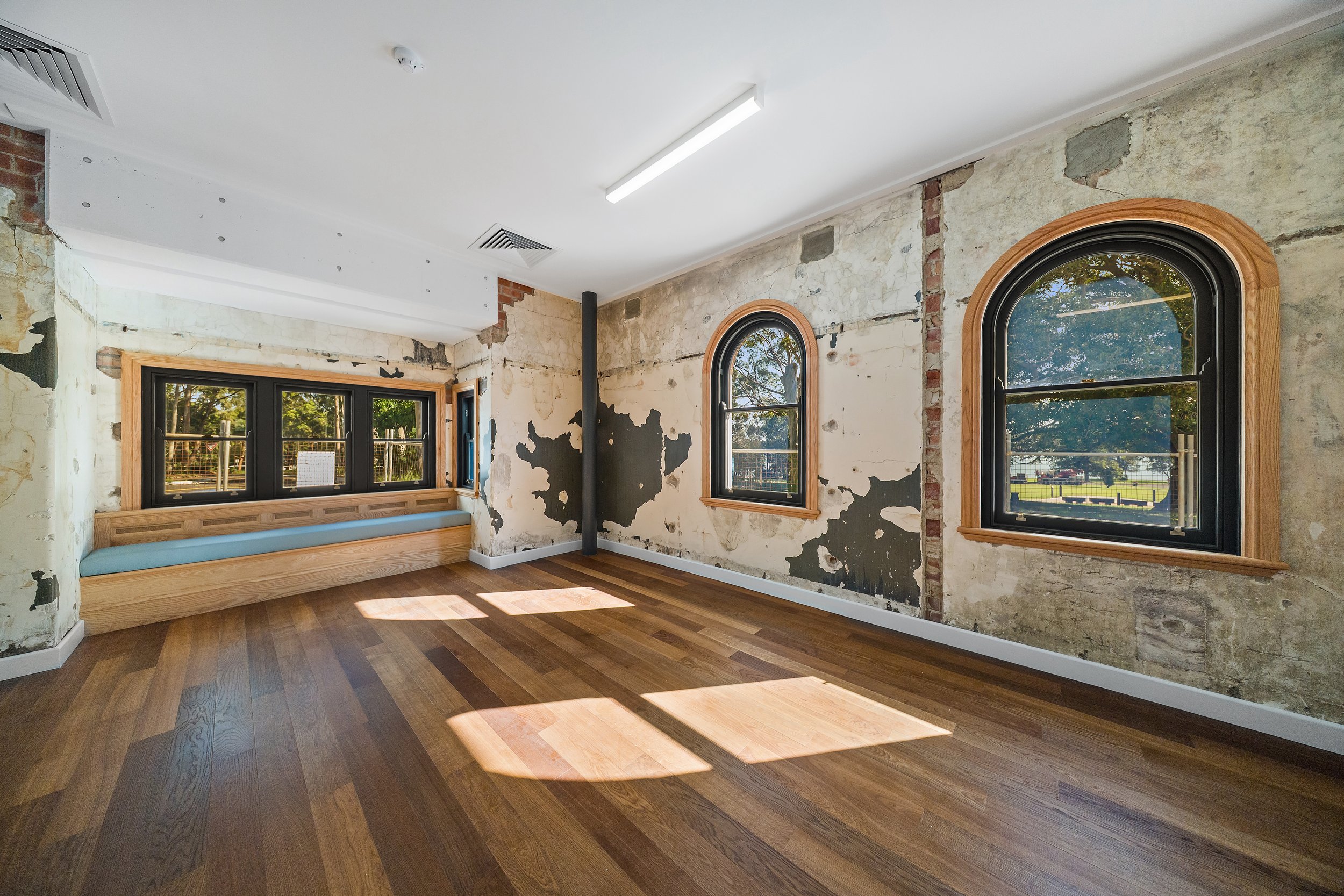 Interior of a room under renovation with exposed brick and plaster walls, wood flooring, and arched windows showing outdoor trees and grass.