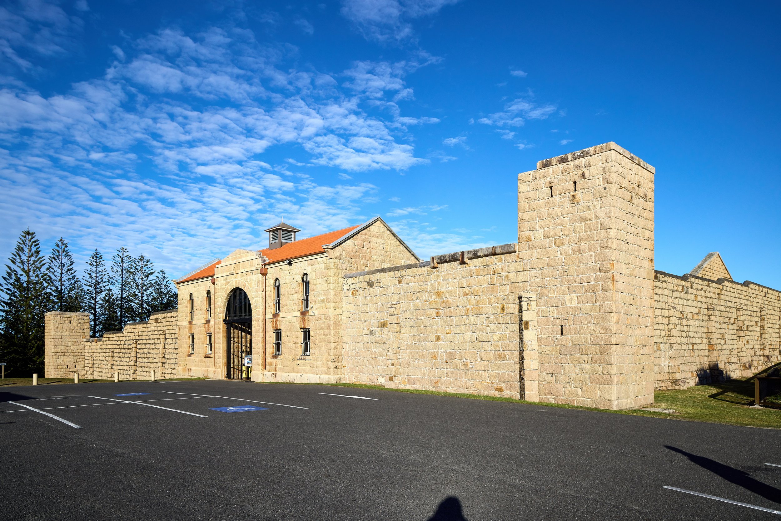 A stone building with a red-tiled roof and a chimney, surrounded by a stone wall, under a blue sky with scattered clouds.