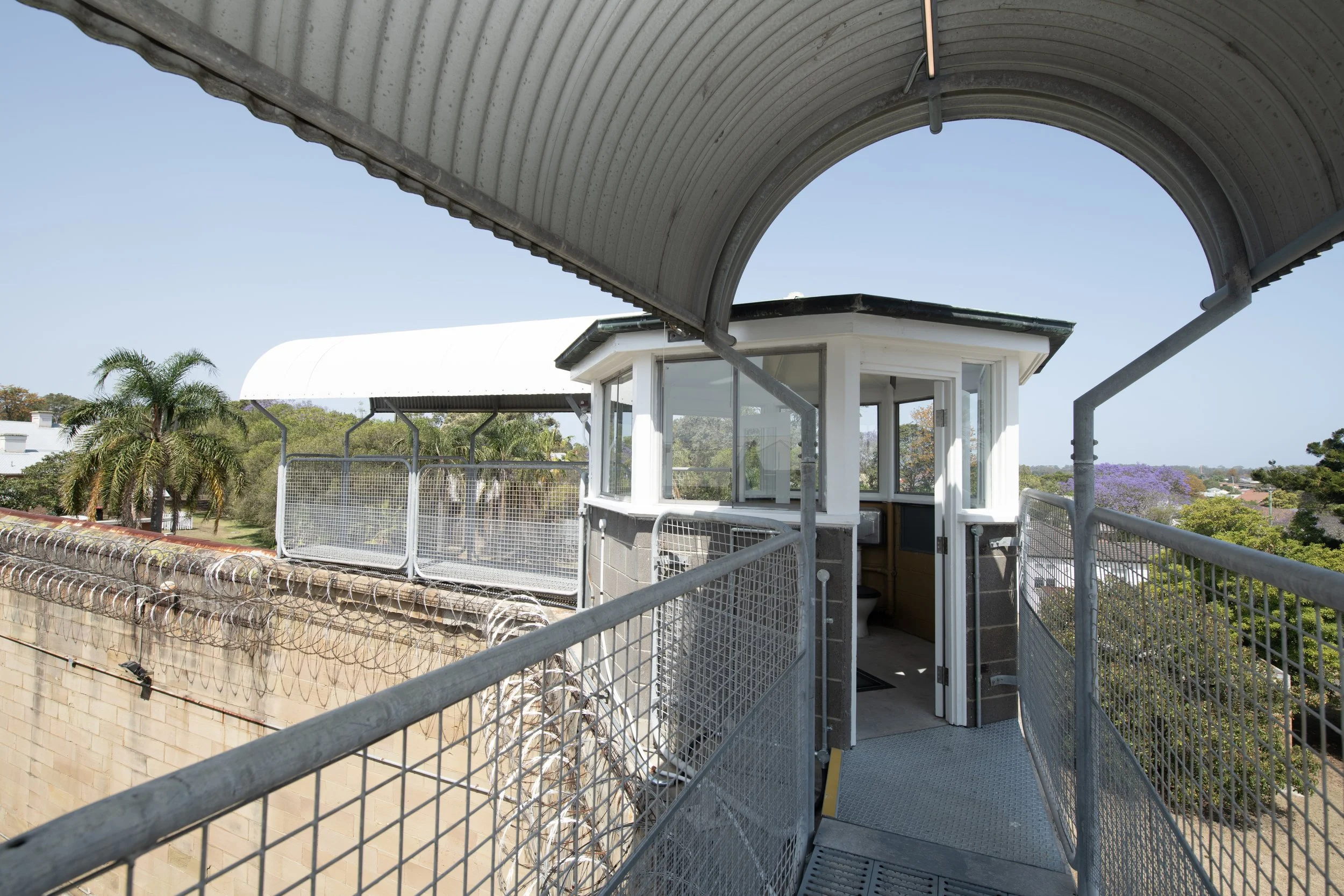 View of a rooftop observation deck with a security booth, surrounded by metal railings, with trees and buildings in the background under a clear blue sky.